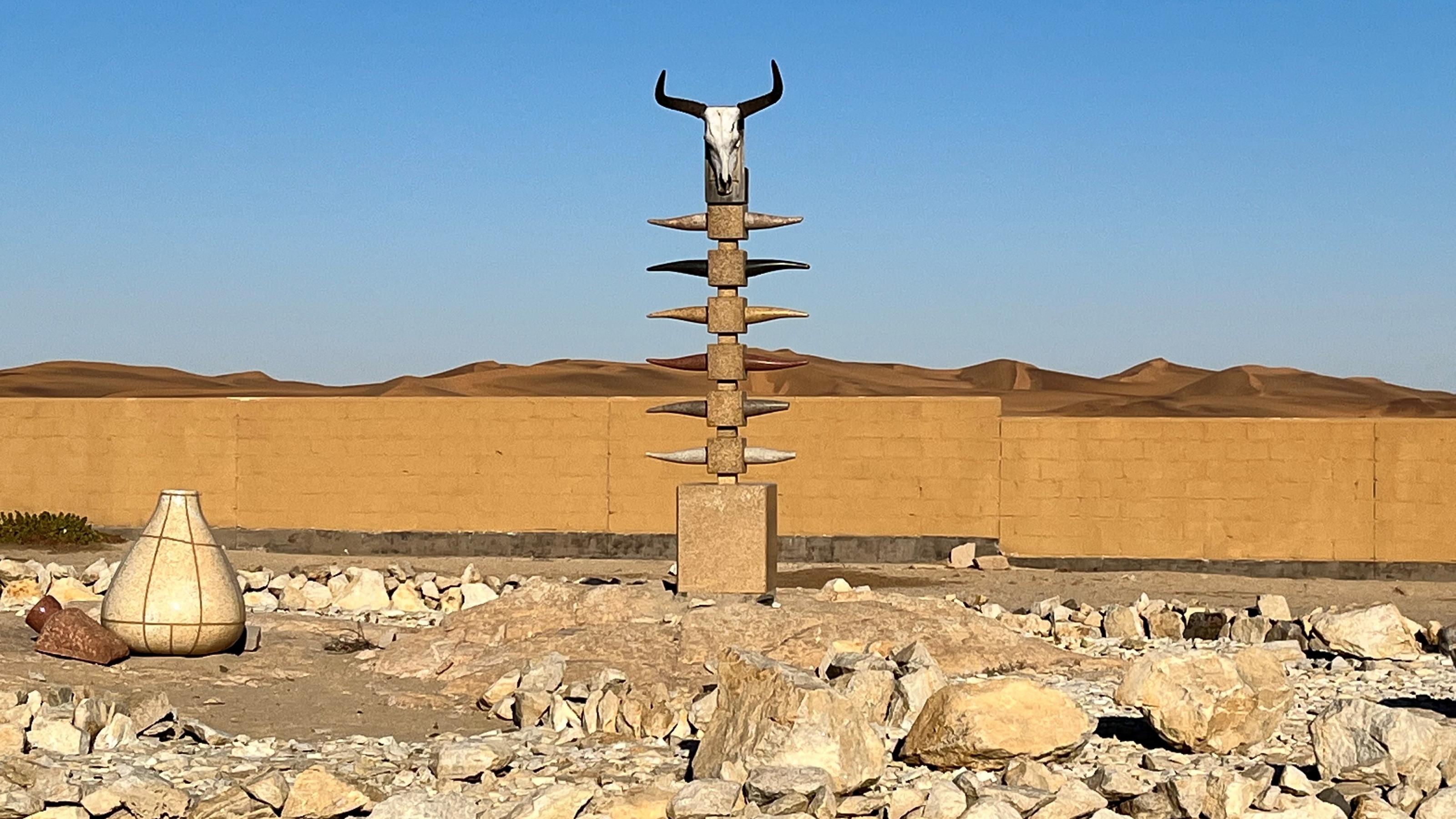 In einer Wüstenlandschaft ragt eine Skulptur in den Himmel, Ganz oben ein Rinderschädel, an der Seite symbolische Hörner, umgeben von Steinen.