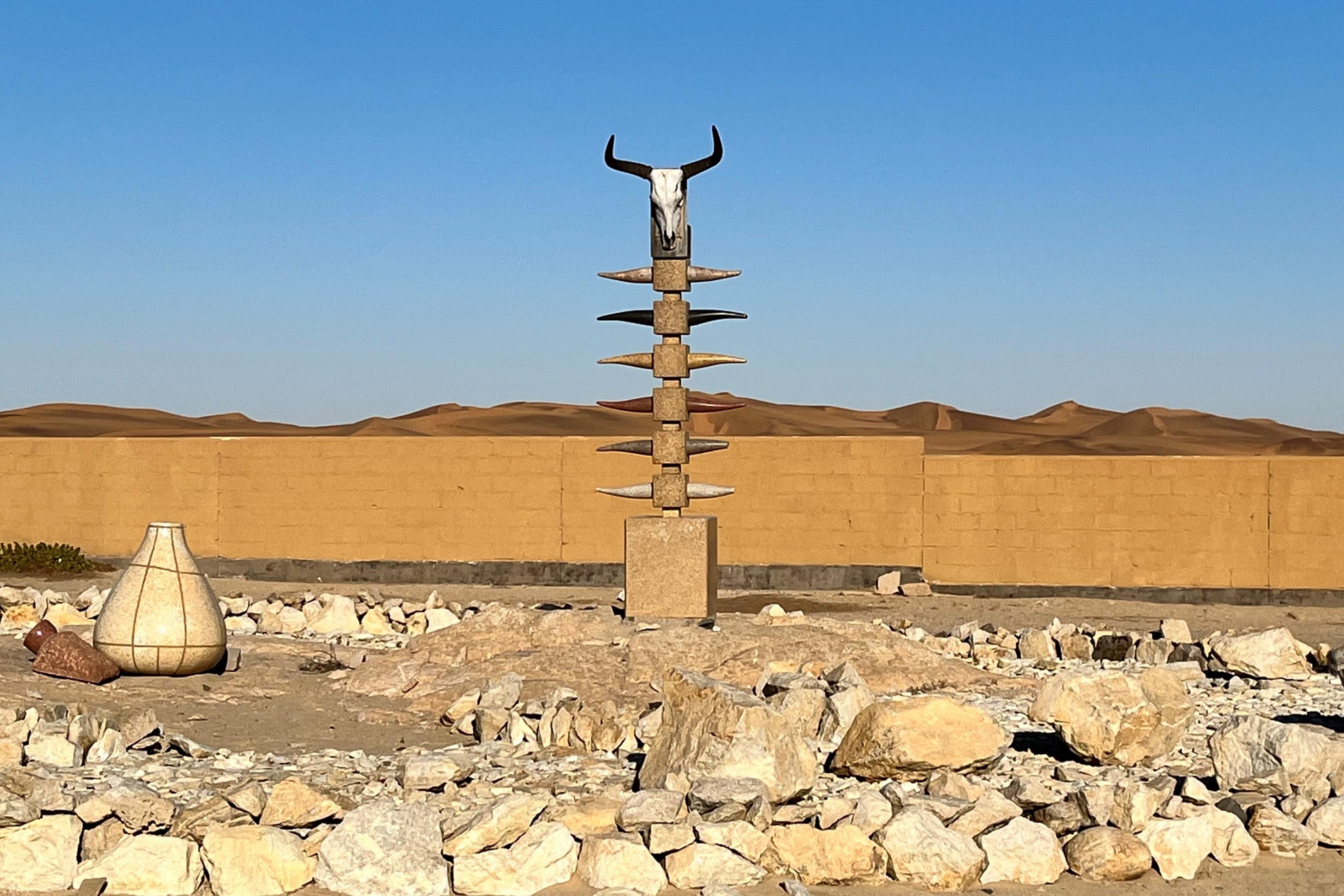 In einer Wüstenlandschaft ragt eine Skulptur in den Himmel, Ganz oben ein Rinderschädel, an der Seite symbolische Hörner, umgeben von Steinen.