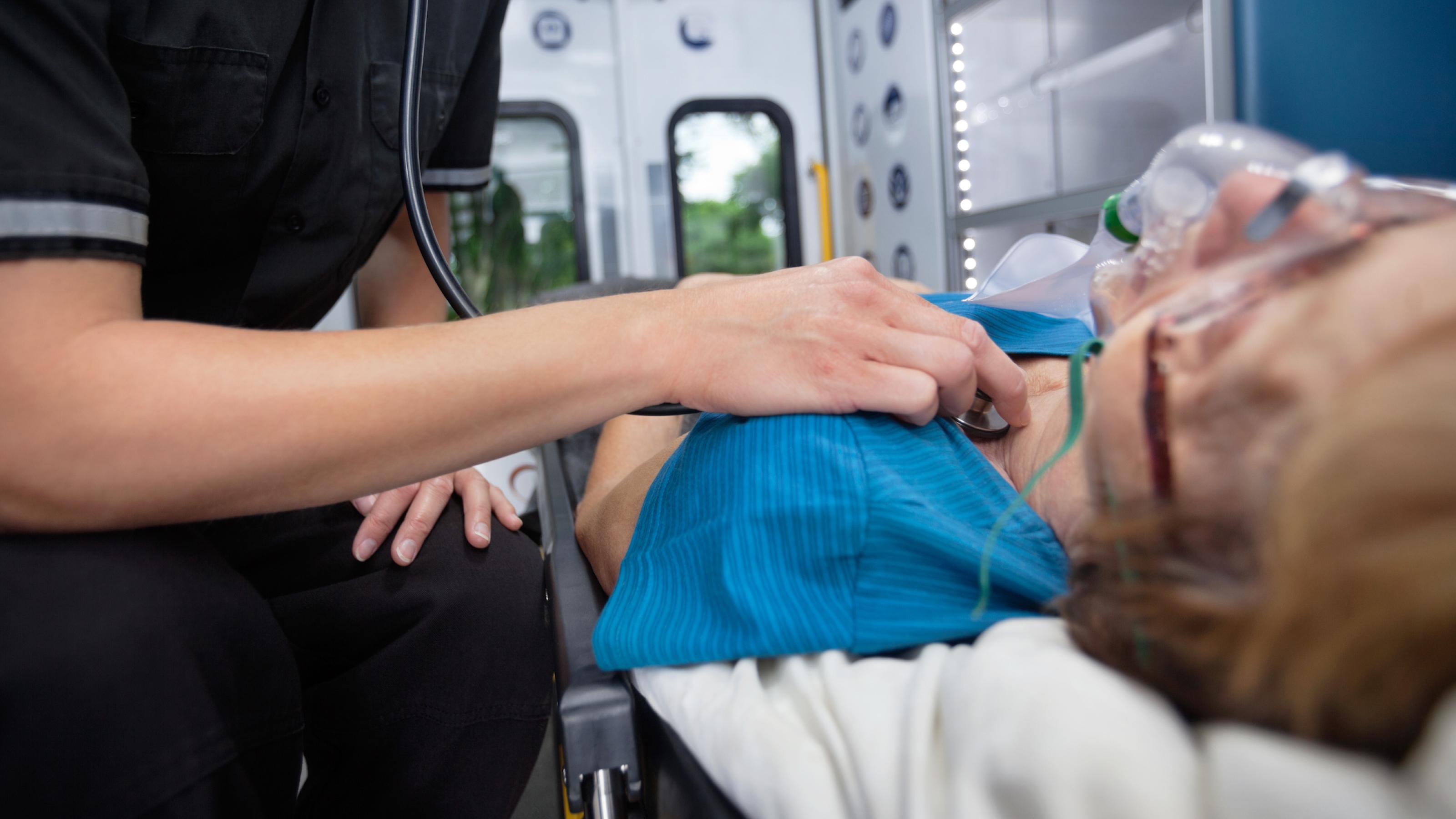 Eine Frau liegt mit einer Sauerstoffmaske auf dem Gesicht auf einer Trage. Eine Person in Uniform misst gerade mit dem Stethoskop ihren Puls. Im Hintergrund sind die Türen eines Krankenwagens und durch die Fenster hindurch verschwommen eine Landschaft zu sehen.