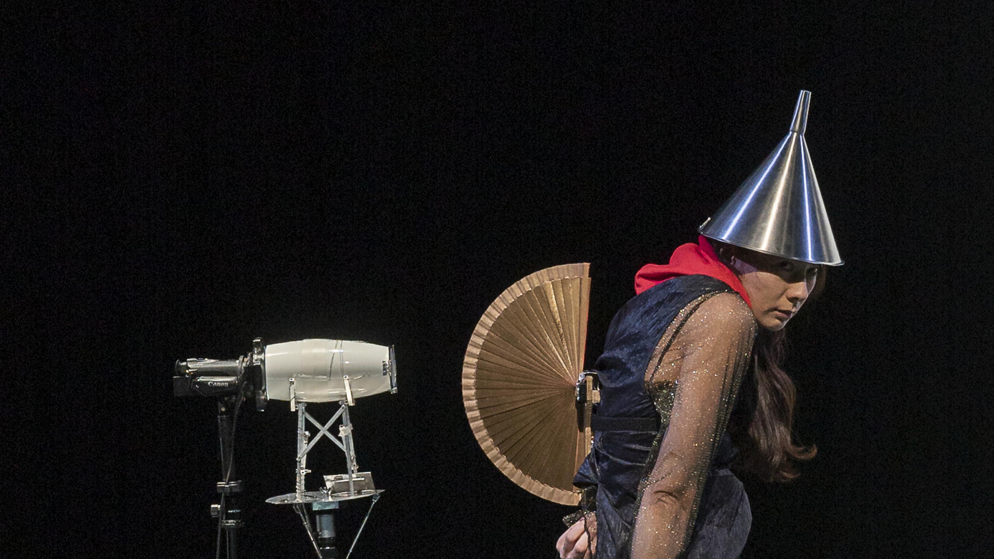 Die Darstellerin Elisabeth Hofmann steht auf der Bühne, als sei sie eine Figur aus einem Bild von Hieronymus Bosch. Sie trägt einen Trichter auf dem Kopf und einen geöffneten Fächer auf dem Rücken, wie eine Eidechse mit aufgeplustertem Kamm.