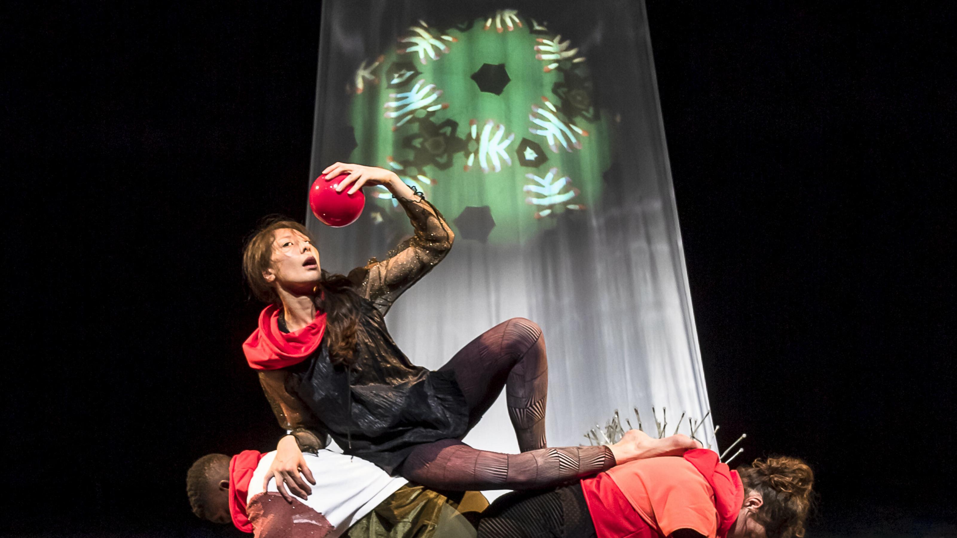 Ein Bild wie aus der Renaissance: Auf dem Rücken von zwei Darsteller:innen hält sich eine weitere Darstellerin einen roten Ball wie eine süße Frucht über ihren geöffneten Mund hält. Im Hintergrund ein weißer Vorhang mit der grünen Projektion eines Kaleidoskops.