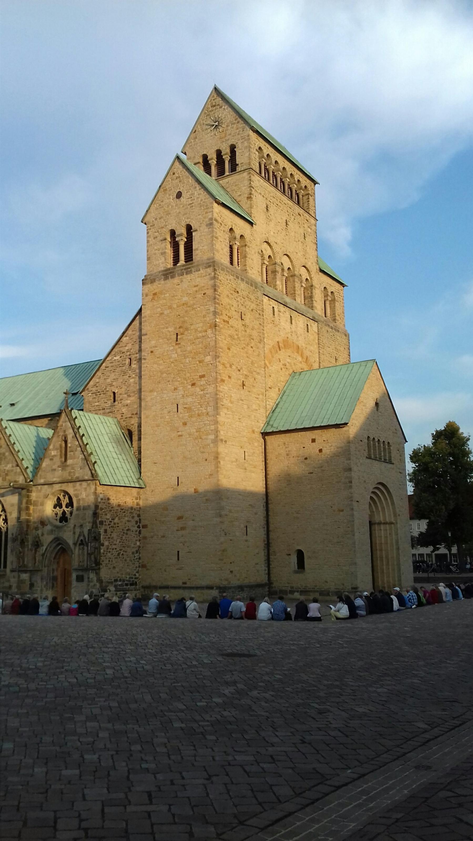 Menschen sitzen auf dem Domhof am Mariendom in Hildesheim und hören einem Glockenkonzert zu.