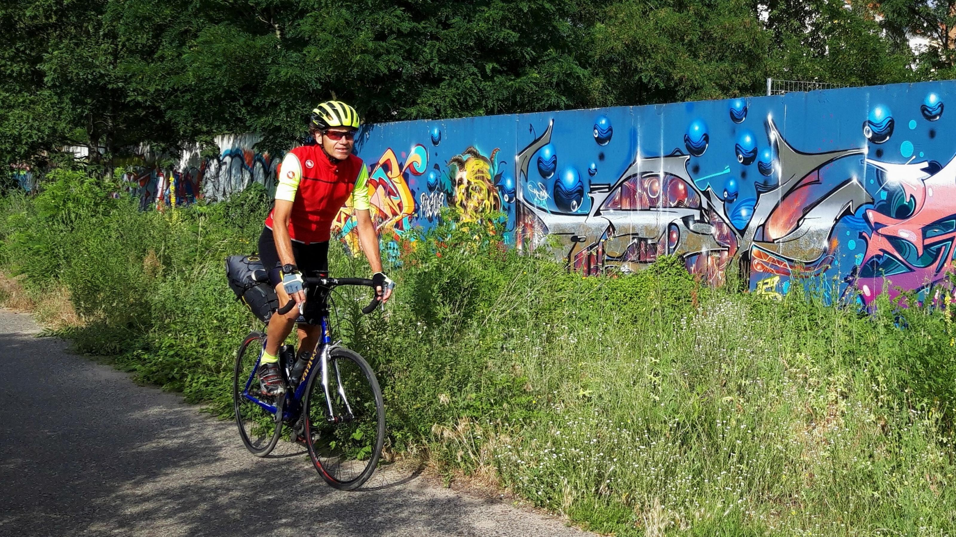 Der gelb behelmte Radfahrer fährt auf seinem Rennrad entlang eines Grünstreifens, hinter dem eine von der Grundfarbe blaue, aber quirlig bunt bemalte und besprühte Mauer aufragt.