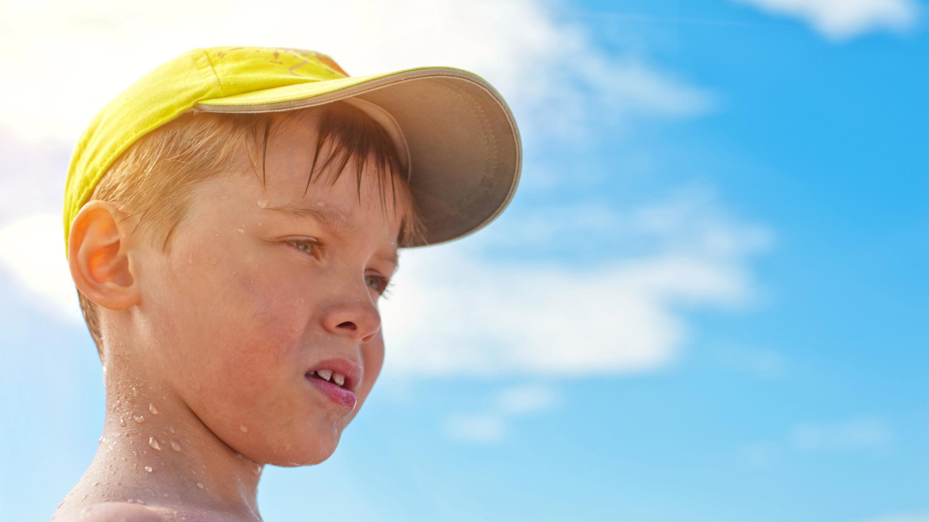 Halbnahe: Ein etwa 6-jähriger Junge mit heller Haut und blonden Haaren im Profil, schaut nach rechts. Er trägt ein gelbes Käppi, Schweißperlen im Gesicht. Im Hintergrund in der Unschärfe Sonne und wenige Wolken.