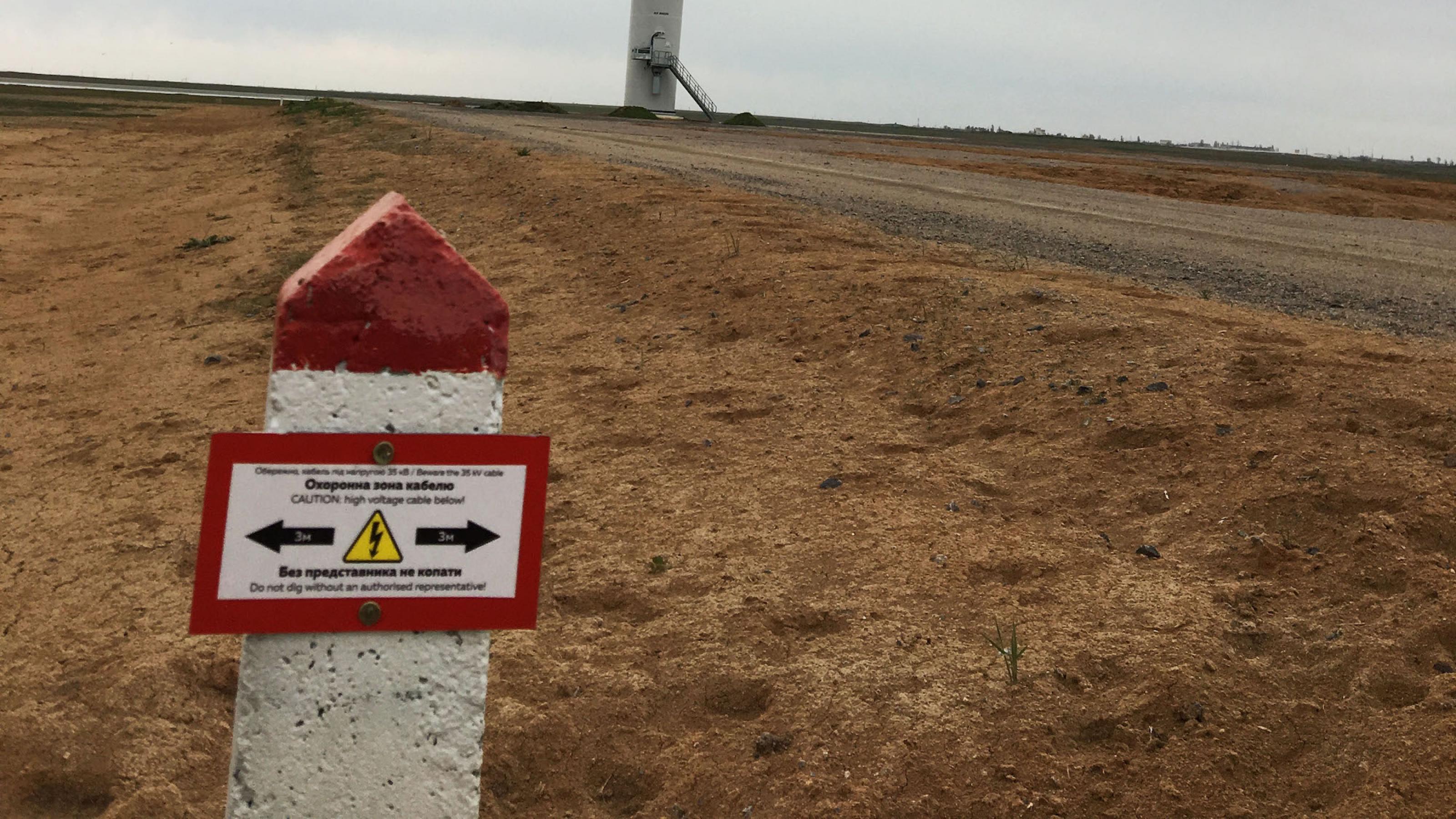 Im Vordergrund ein rot-weißer Warnstein, auf dem vor Hochspannung gewarnt wird. im Hintergrund ist der Fuß einer Windkraftanlage zu erkennen.