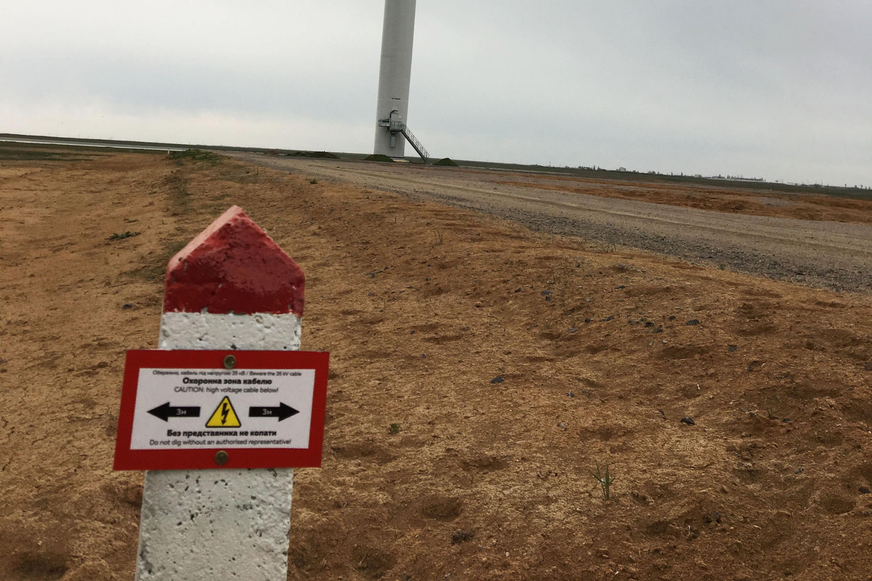 Im Vordergrund ein rot-weißer Warnstein, auf dem vor Hochspannung gewarnt wird. im Hintergrund ist der Fuß einer Windkraftanlage zu erkennen.