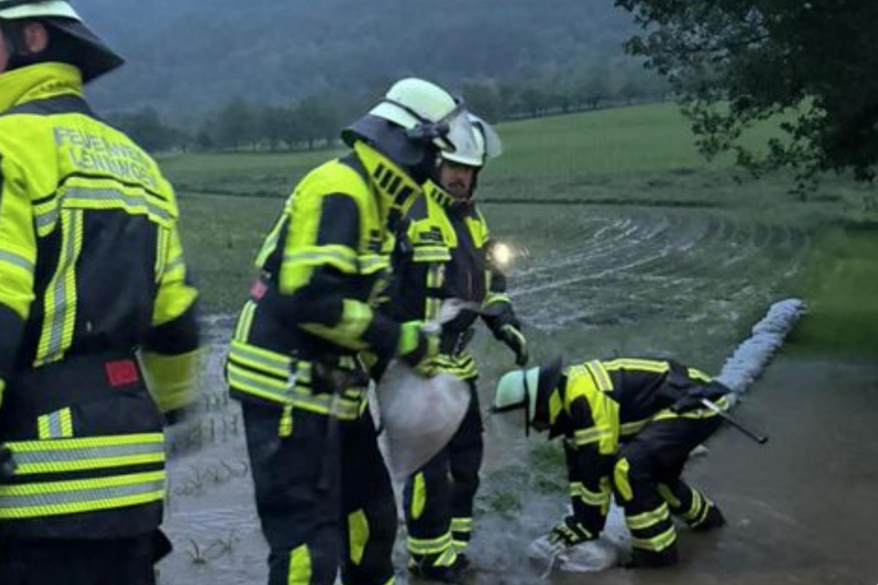 Das Bild zeigt eine Kette junger Männer in reflektierender Schutzkleidung und mit Helmen, die Sandsäcke am Rande eines Feldes auslegen. Der Himmel im Hintergrund ist grau und bedrohlich.