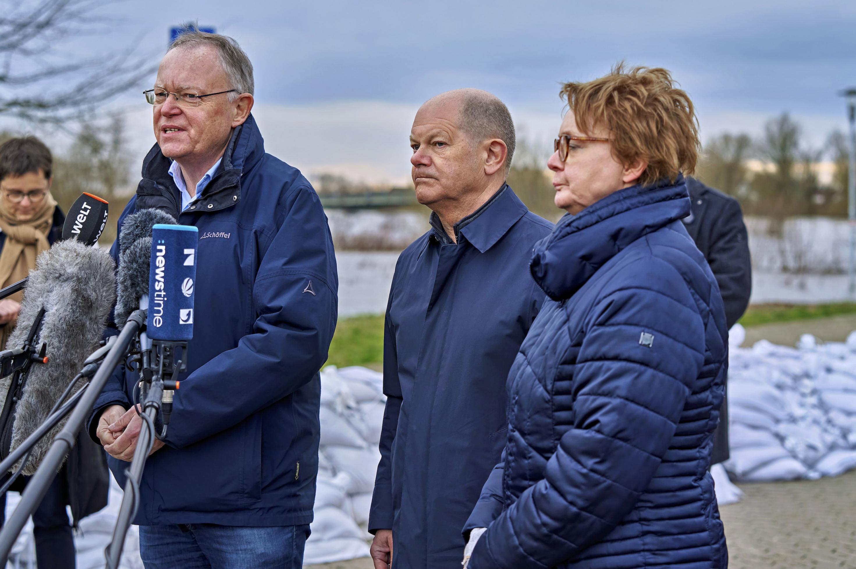 Die drei Personen schauen ernst in Richtung Journalisten, hinter ihnen weiße Sandsäcke zum Schutz des Deichs.