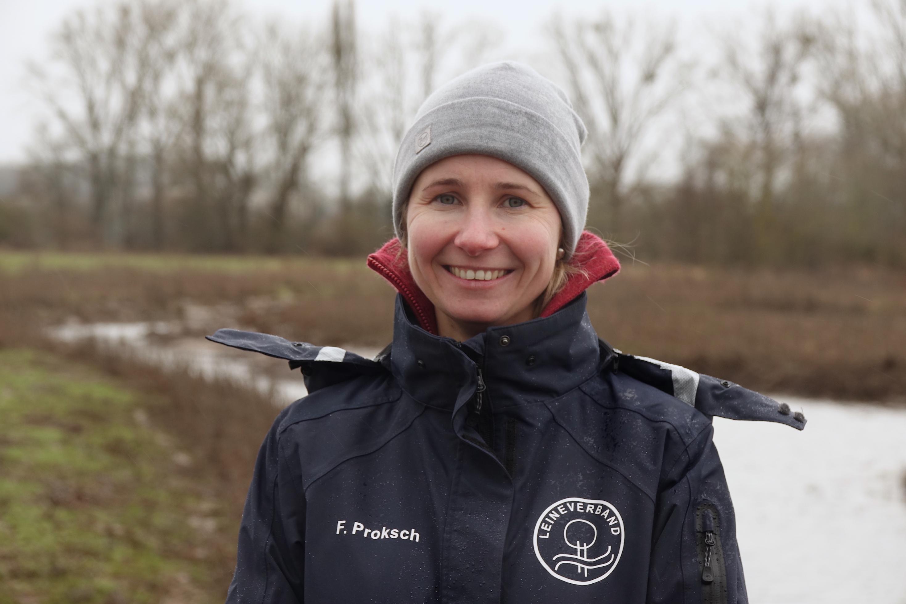 Die junge Frau schaut lächelnd in die Kamera. Sie steht vor einer überschwemmten Landschaft.