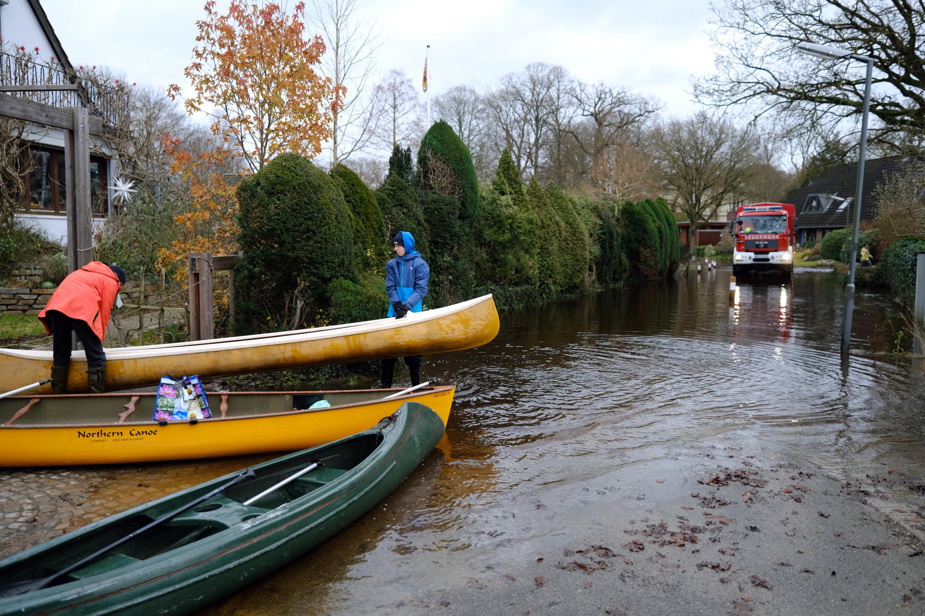 Zwei Jugendliche tragen ein Kanu an einer überfluteten Straße in Borgfeld bei Bremen. Hier ist die Wümme über die Ufer getreten. Die Anwohnerinnen und Anwohner können ihre Häuser nur noch mit Booten erreichen.