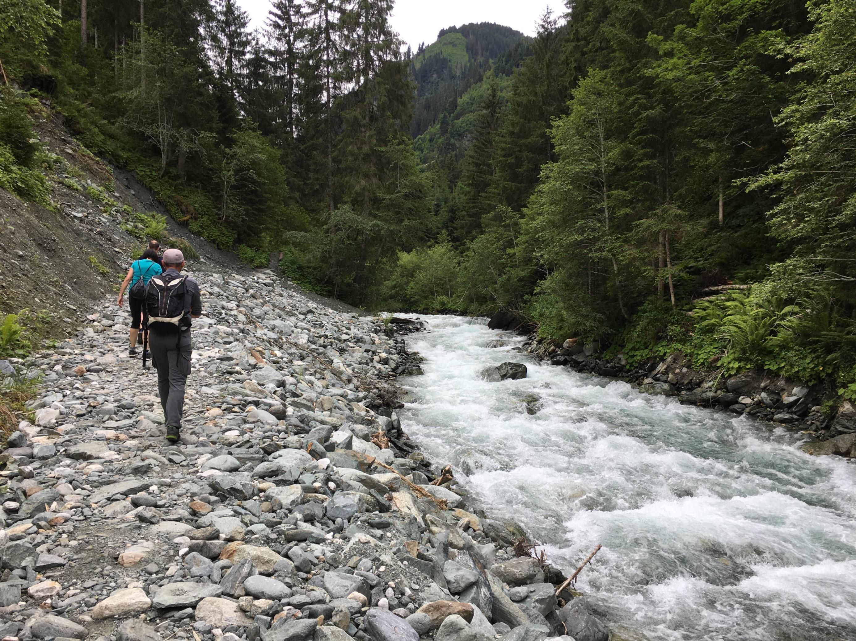 Gebirgsbach, links ein steiniger Wanderweg mit Wanderern.