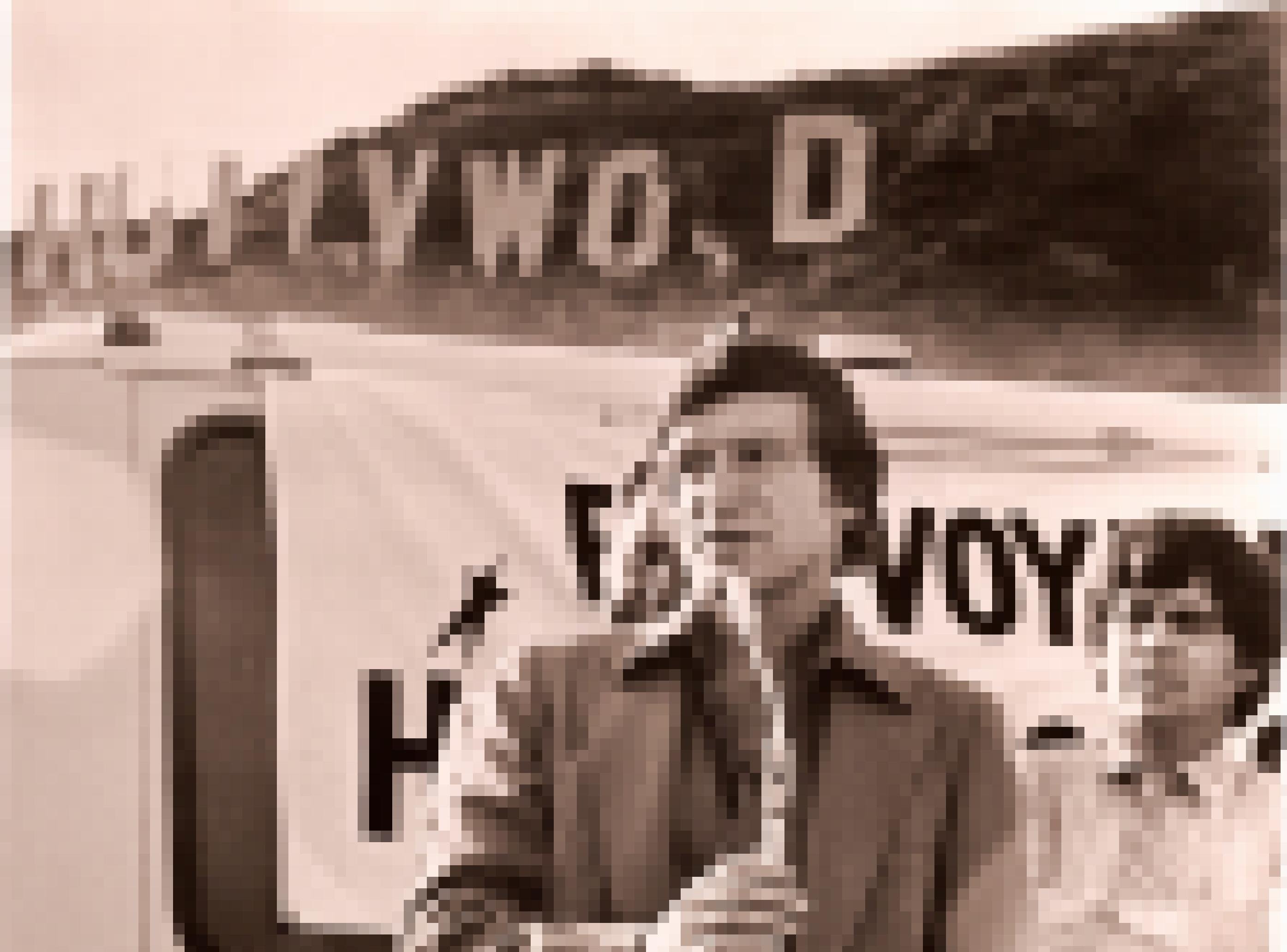 schwarz-weiss Foto von Playboy Herausgeber Hugh Hefner Ende der 70er. Er steht im Anzug mit offenem Kragen an einem Mikrofon. Im Hintergrund das Hollywood Zeichen, von dem eine Buchstaben verrottet sind