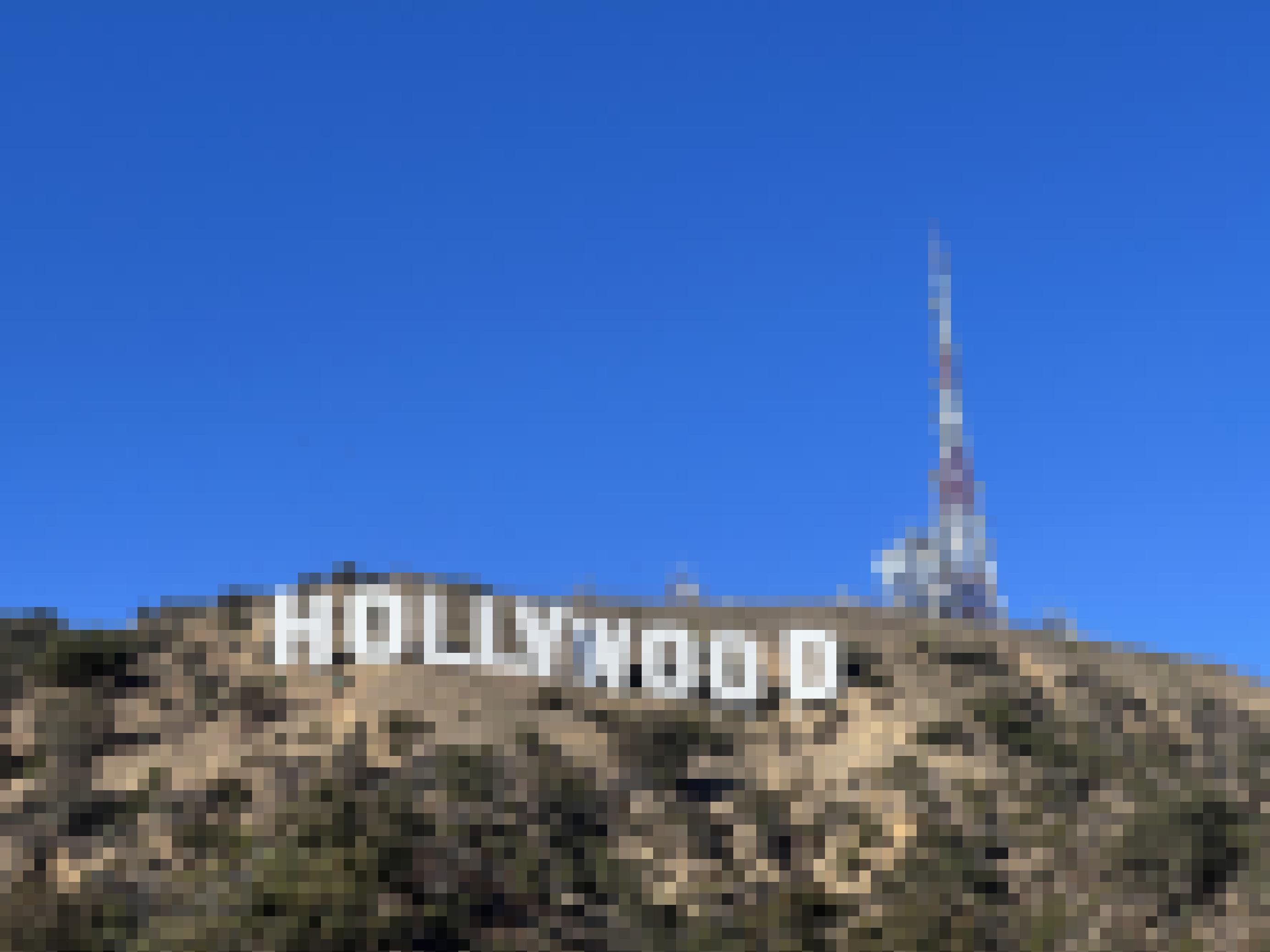 Der strahlend weiße Hollywood Schriftzug in den Hügeln, im Hintergrund die Türme eines Elektrizitätswerkes, d arüber viel wolkenloser Himmel.