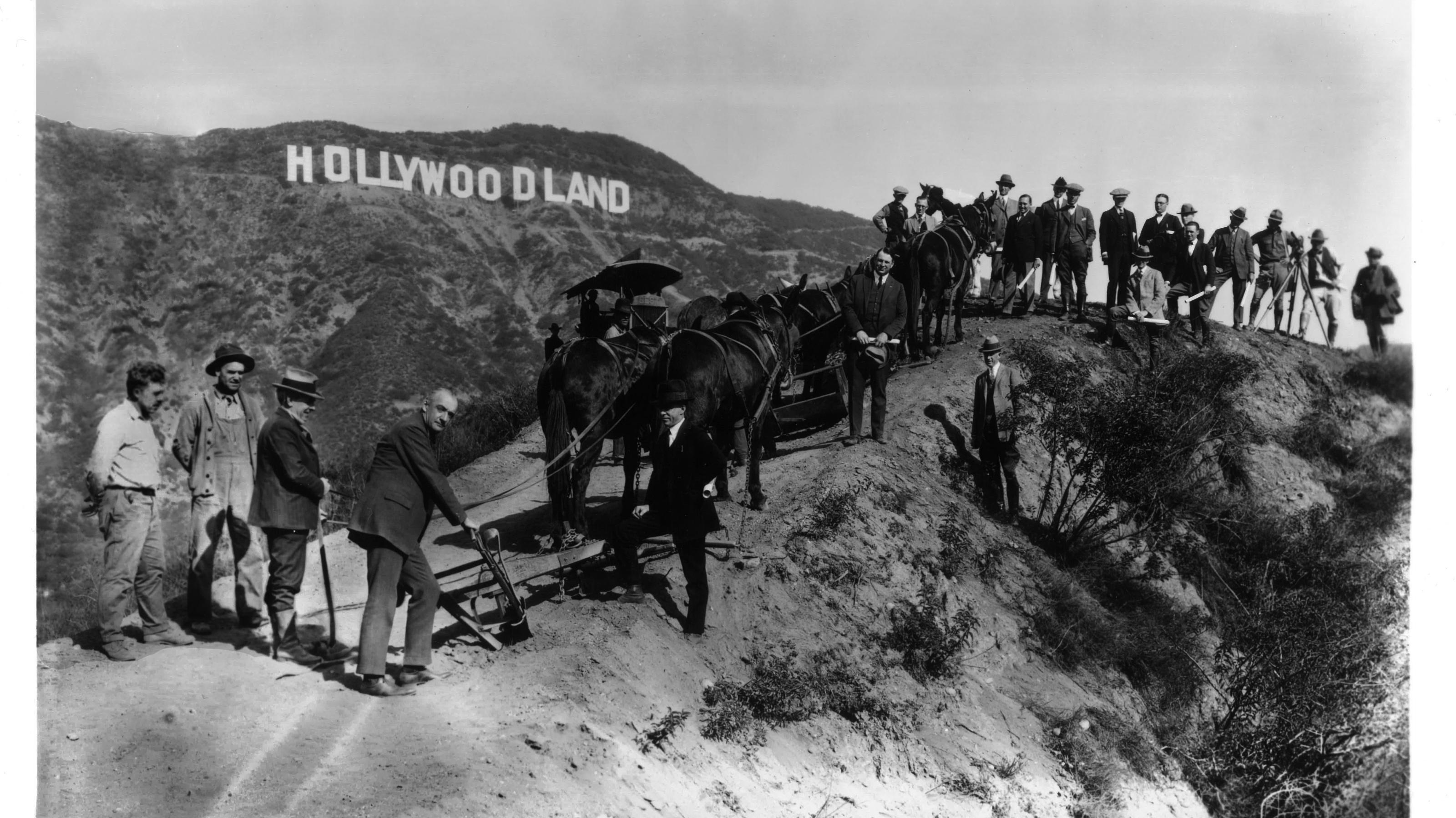 Schwarz-weiß Foto von Männern in den Hügeln vor dem Hollywood Zeichen beim Bau 1923. Damals noch Hollywoodland. Mit Eseln und Dampfmaschinen auf einem unbefestigten Weg.