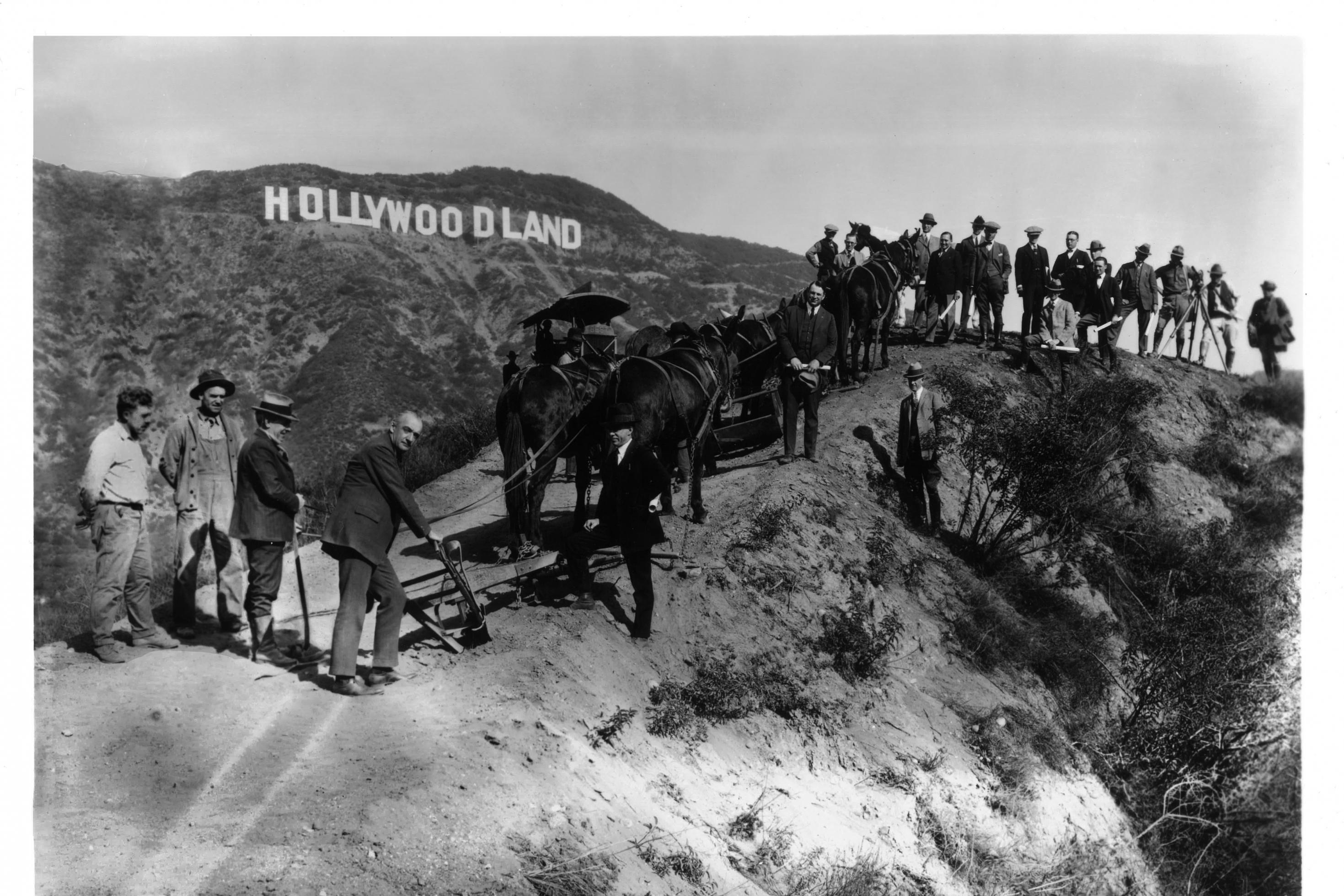 Schwarz-weiß Foto von Männern in den Hügeln vor dem Hollywood Zeichen beim Bau 1923. Damals noch Hollywoodland. Mit Eseln und Dampfmaschinen auf einem unbefestigten Weg.