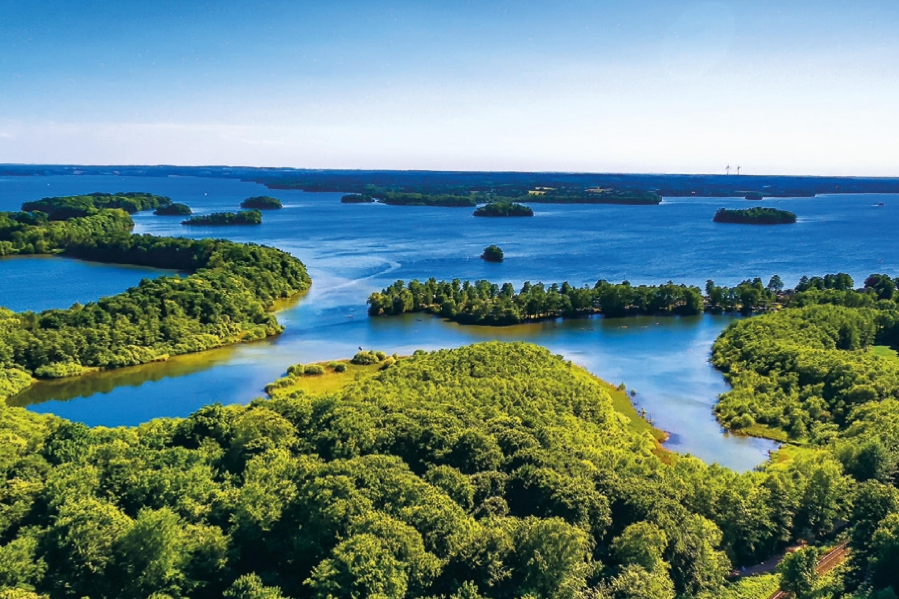 Unter blauem Himmel erstrecken sich zahlreiche tiefblaue Wasserflächen, die von baumreichem Grün umrandet werden.