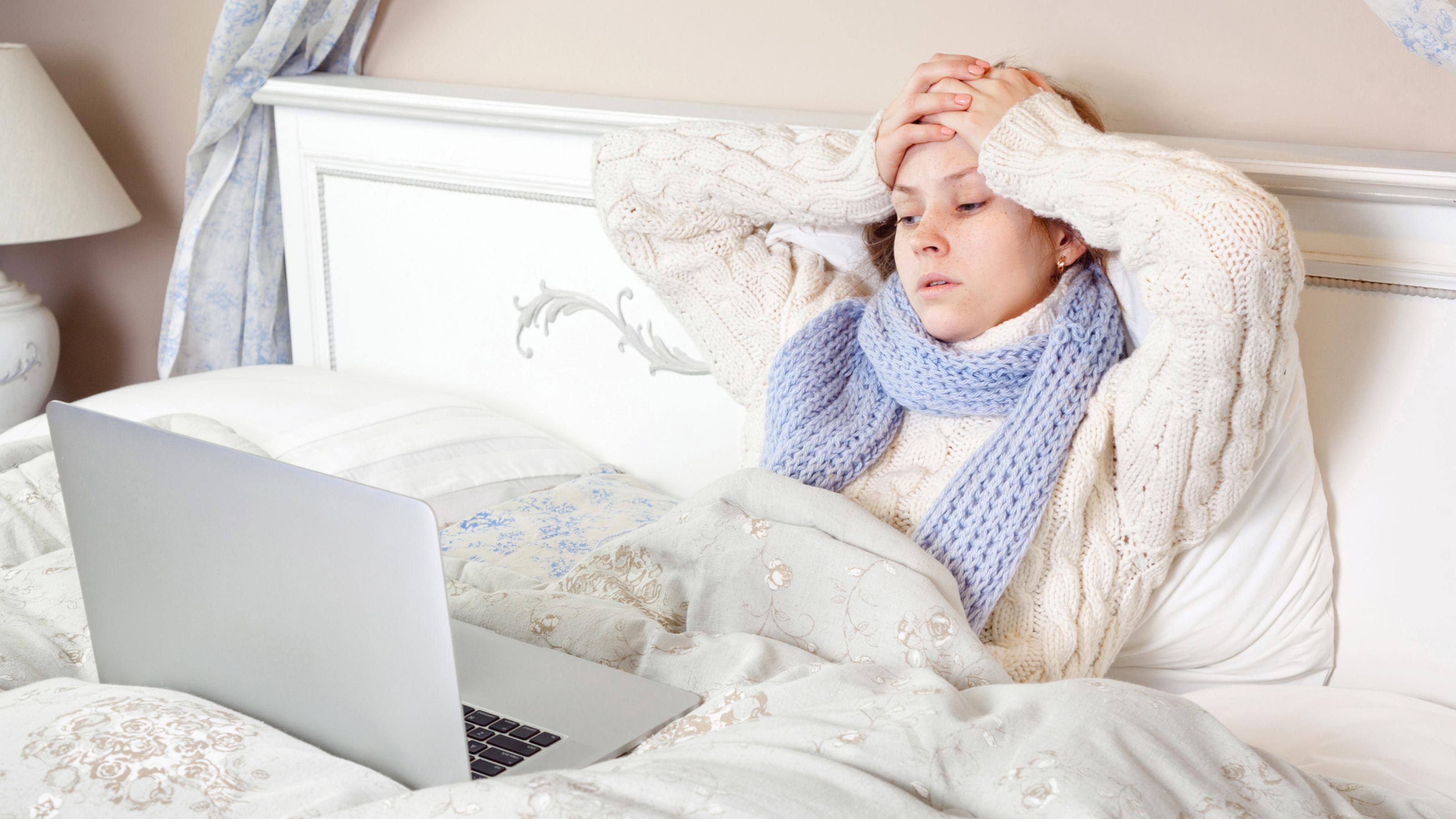 Eine kranke, erschöpft aussehende Frau liegt mit dem Laptop im Bett und versucht, trotz Krankheit ihre Arbeit zu erledigen.