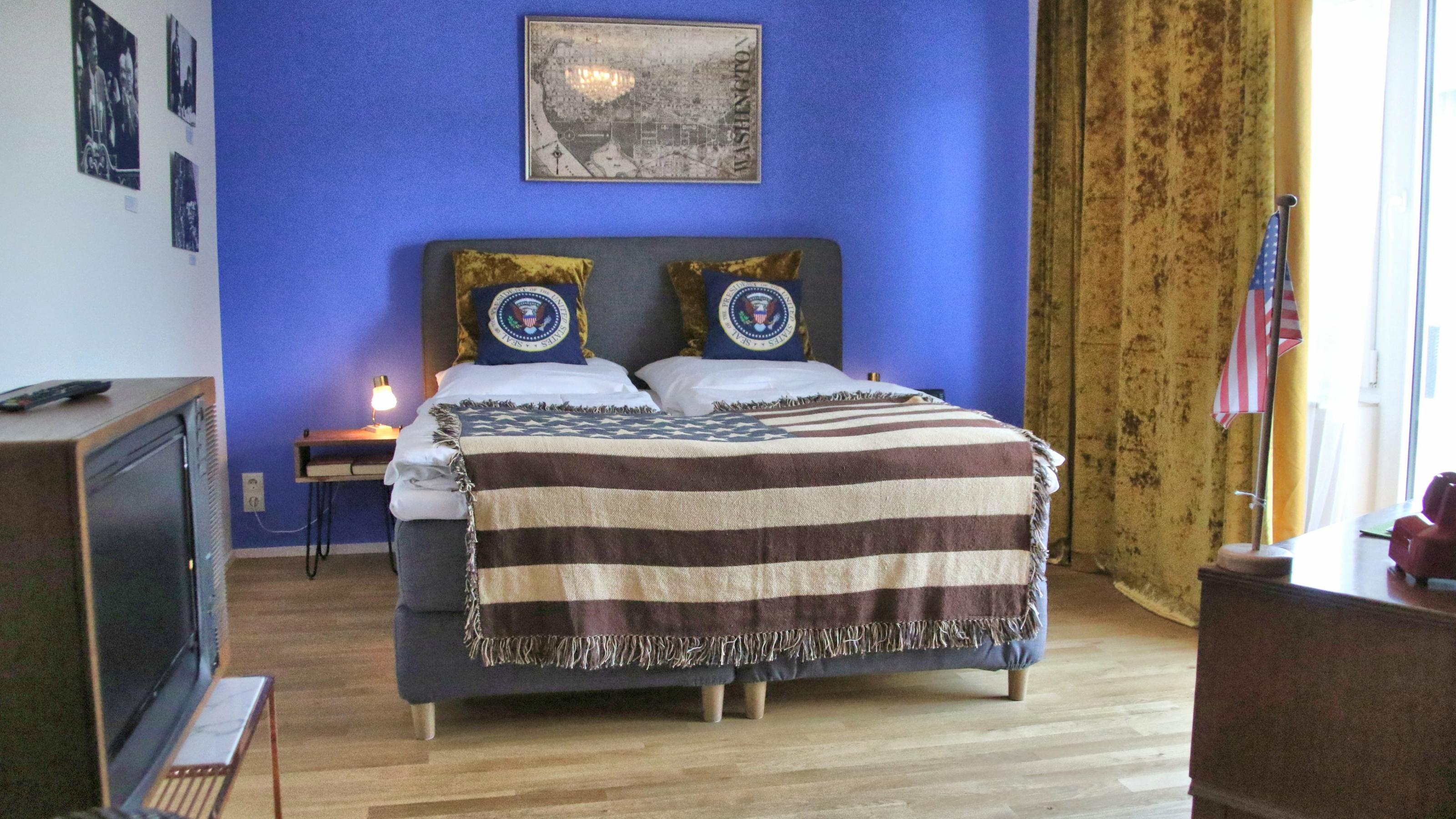 Ein Hotelzimmer mit Bett, auf dem eine Tagesdecke mit den „Stars and Stripes“ der USA liegt. Die Kopfkissen tragen das Präsidenten-Siegel.