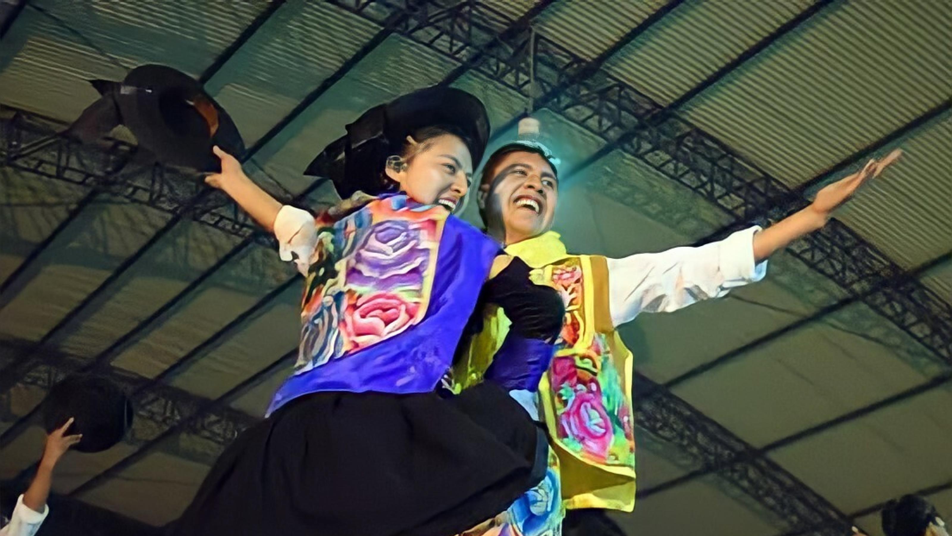Ein junger Mann und eine junge Frau auf einer Bühne. Beide mit farbenfroh bestickten Rock bzw. Weste und schwarzem Hut. Der Mann hält den Hut in der Hand. Blicken lachend von der Bühne herunter.