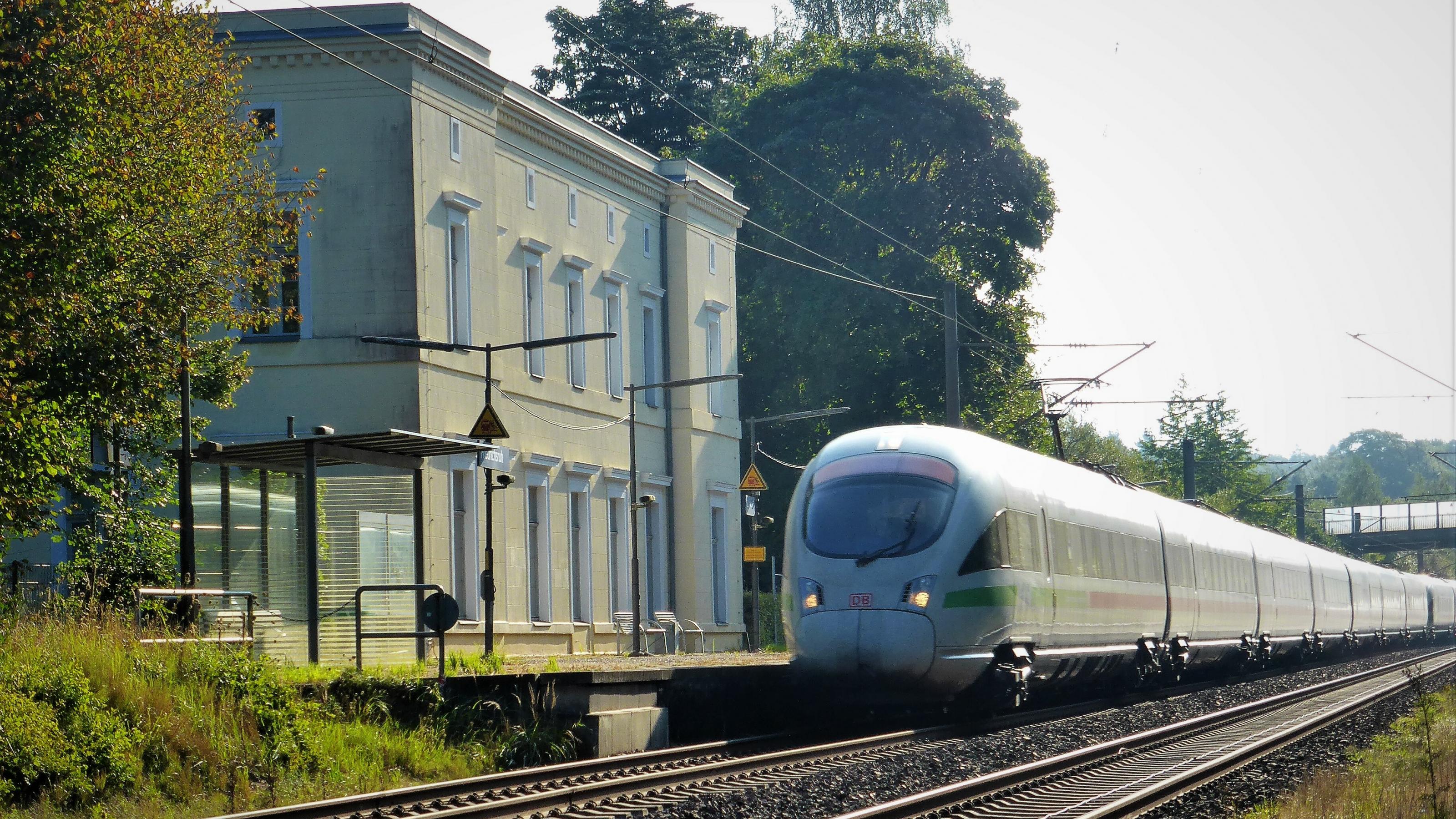 Vor dem klassizistischen, hellbeigen Bahnhofsgebäude liegt ein doppelter Schienenstrang. Auf jenem dem Gebäude nahe Liegenden rauscht ein ICE heran, dessen rundlicher windschnittiger Triebwagen die Bildmitte ausfüllt.