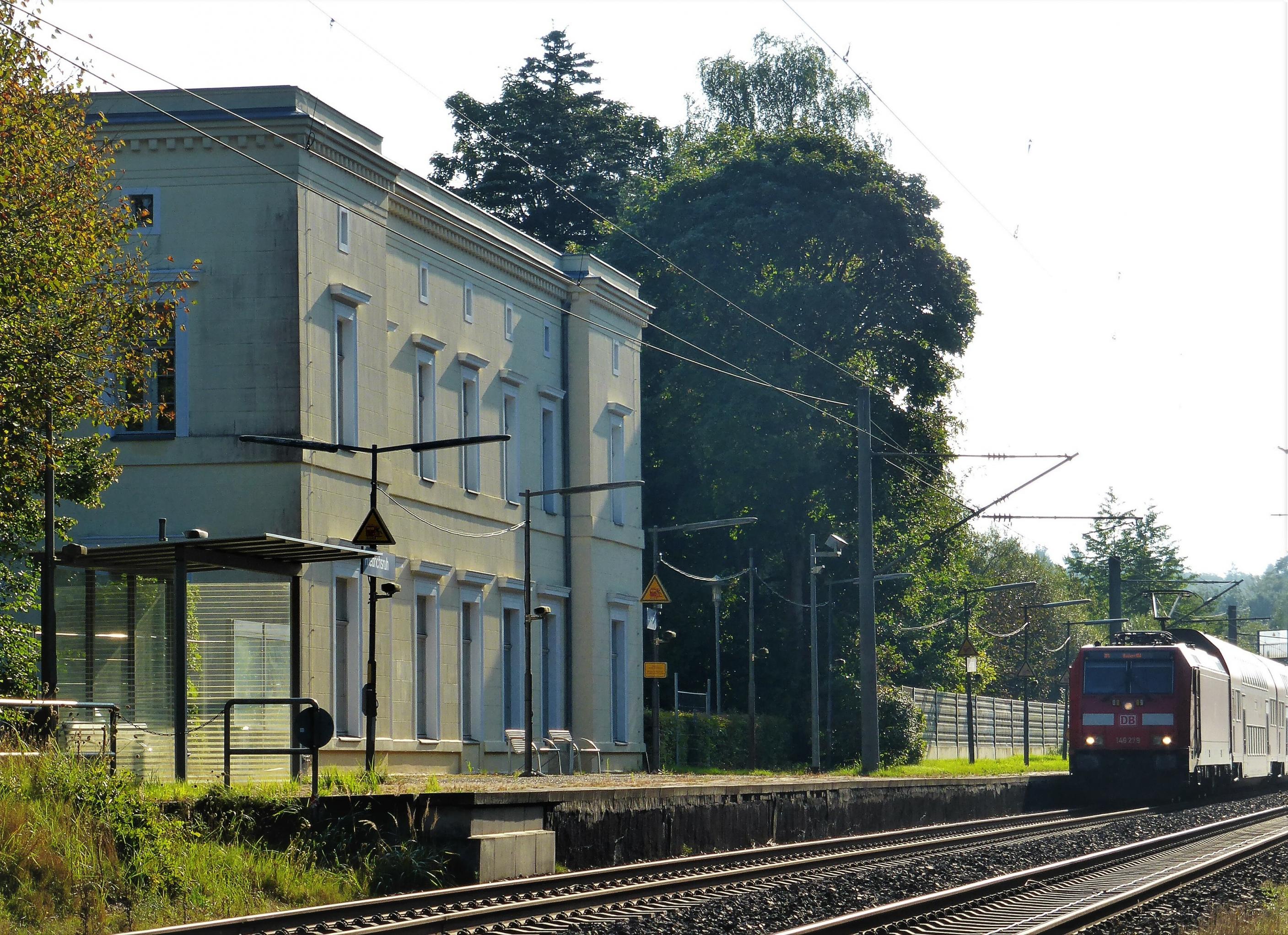 Vor dem klassizistischen, hellbeigen Bahnhofsgebäude liegt ein doppelter Schienenstrang. Auf jenem dem Gebäude nahe Liegenden nähert sich die rote Lok eines Regionalexpress.