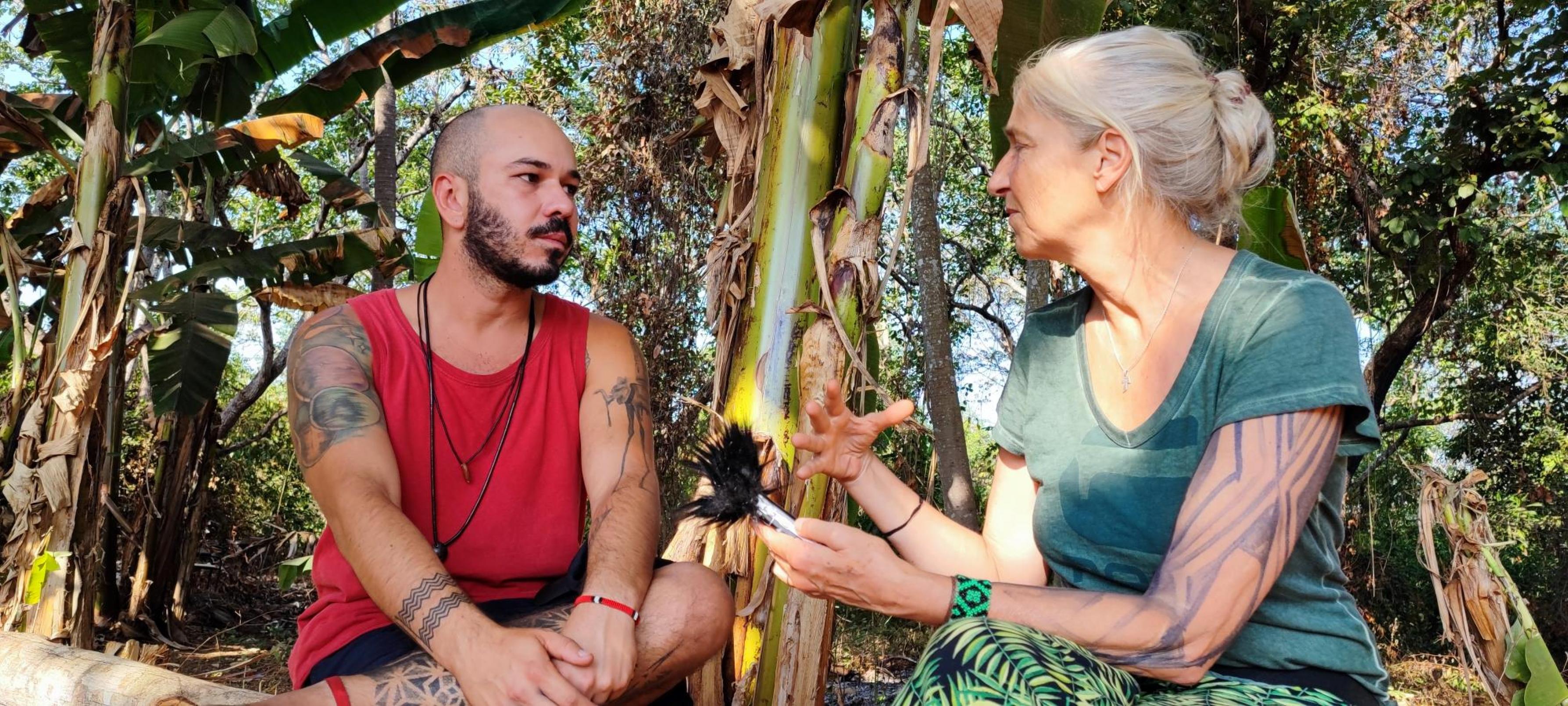 Ein Mann mit Tatoos, indigenem Schmuck und sanftem Blick wird von der Autorin interviewt. Beide sitzen am Rand des Dorfes auf einem Baumstamm.