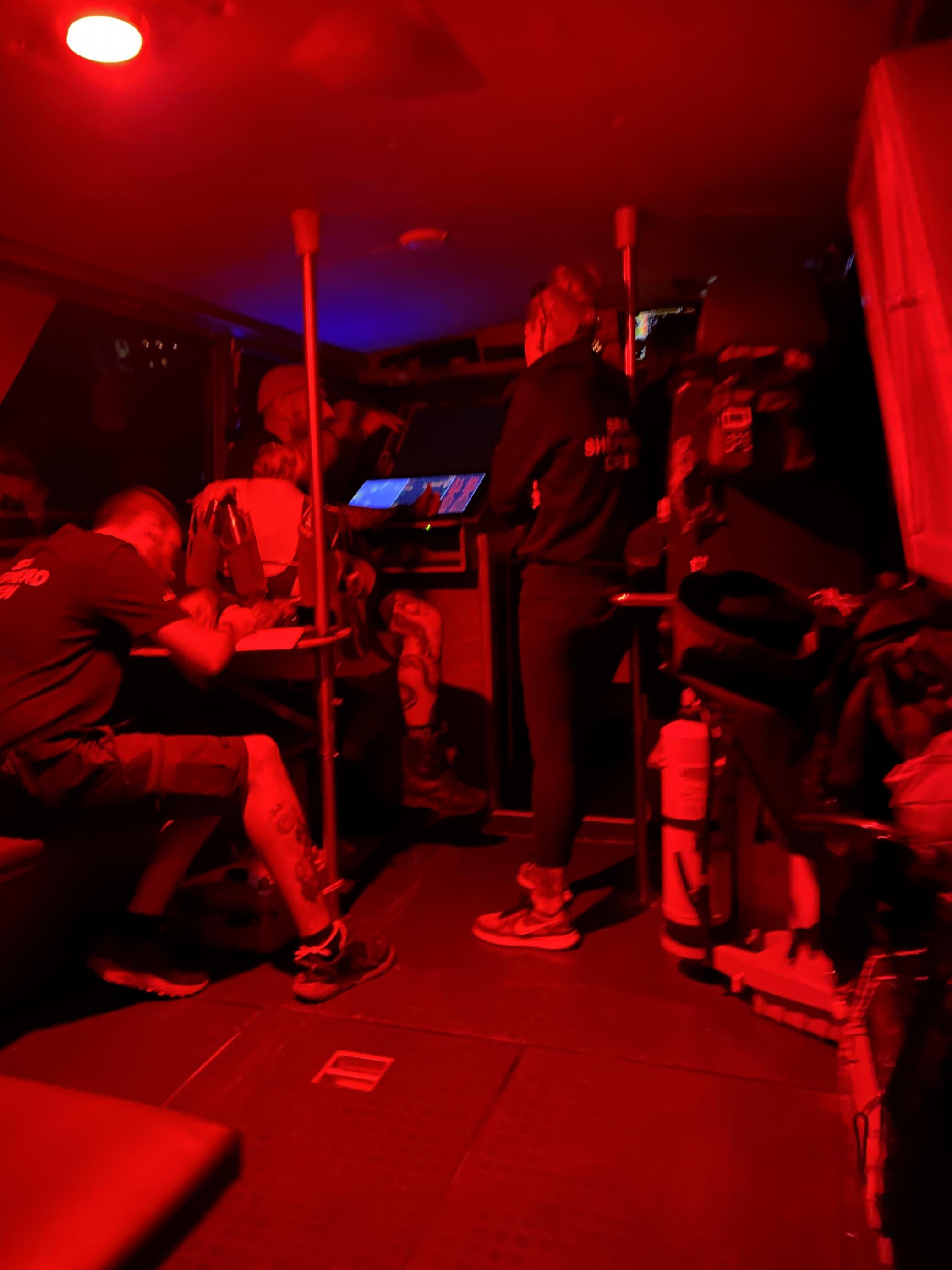Drei Menschen sitzen in der Steuerkajüte eines Bootes, die in rotem Licht ausgeleuchtet ist.