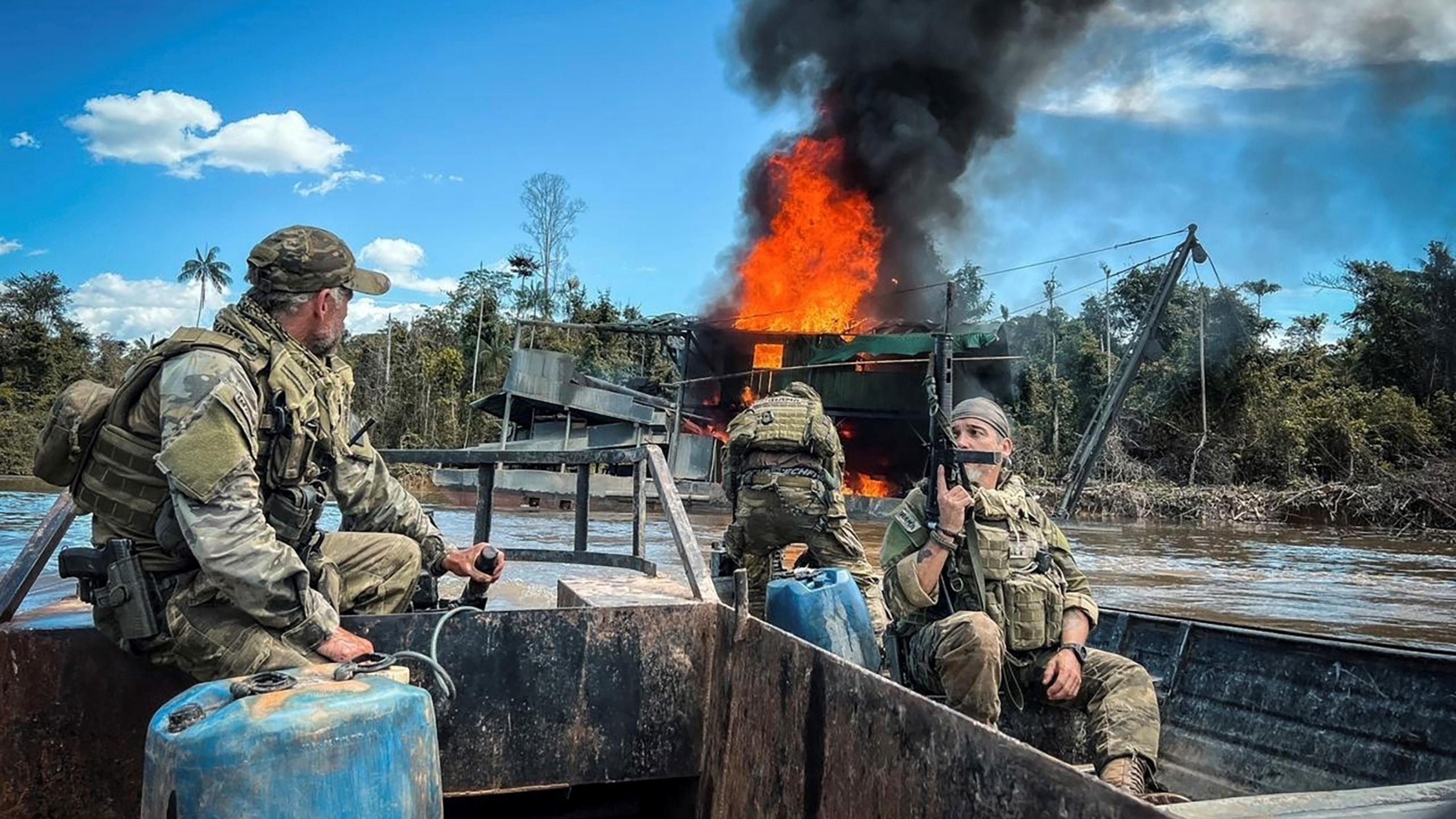 Drei schwer bewaffnete Männer in militärischer Camuflage-Kleidung blicken von ihrem Boot aus auf einen brennenden Bergbaukahn im Hintergrund