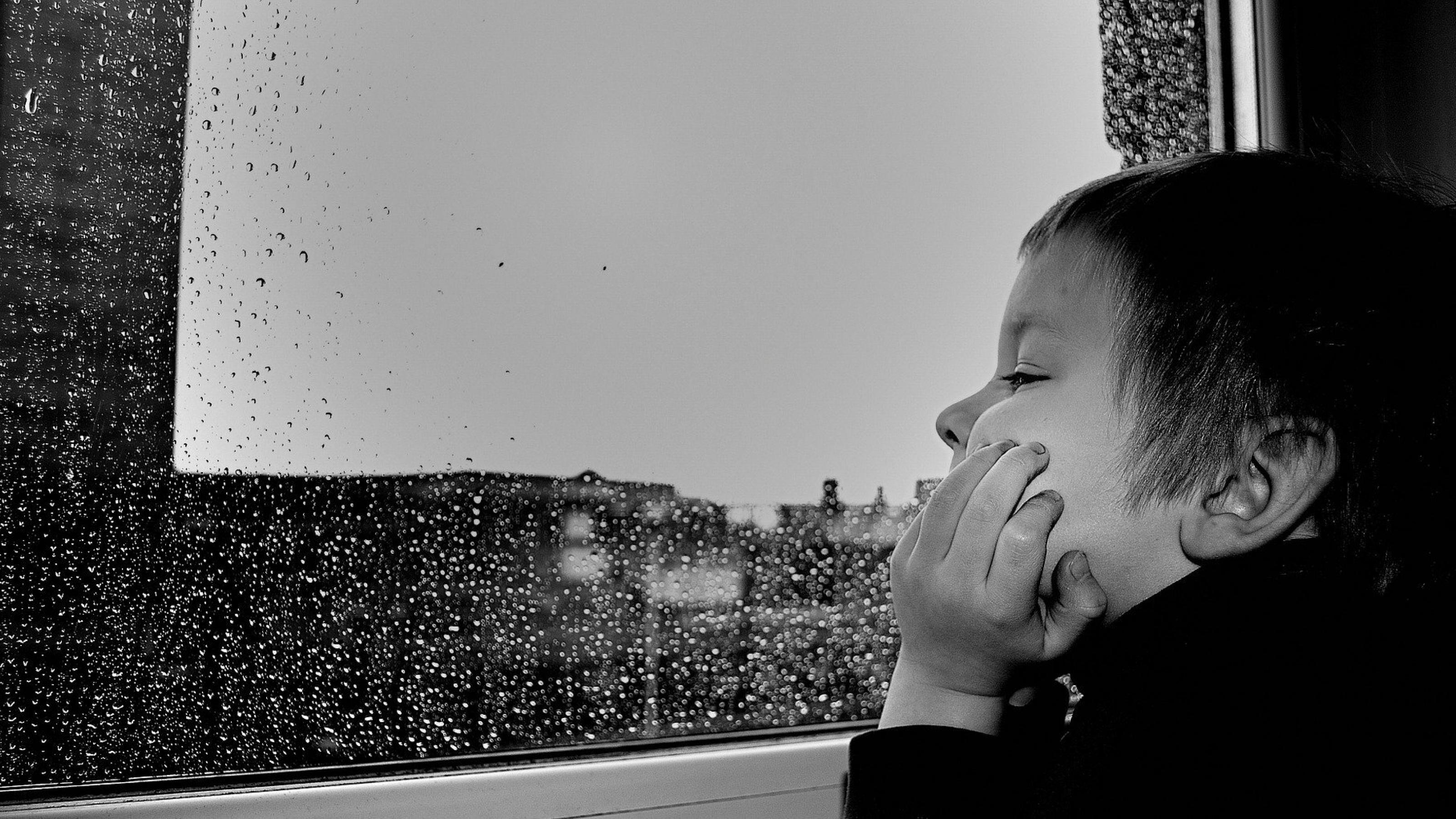 Ein Junge schaut aus einem Fenster. Draußen regnet es, Regentropfen auf der Scheibe.
