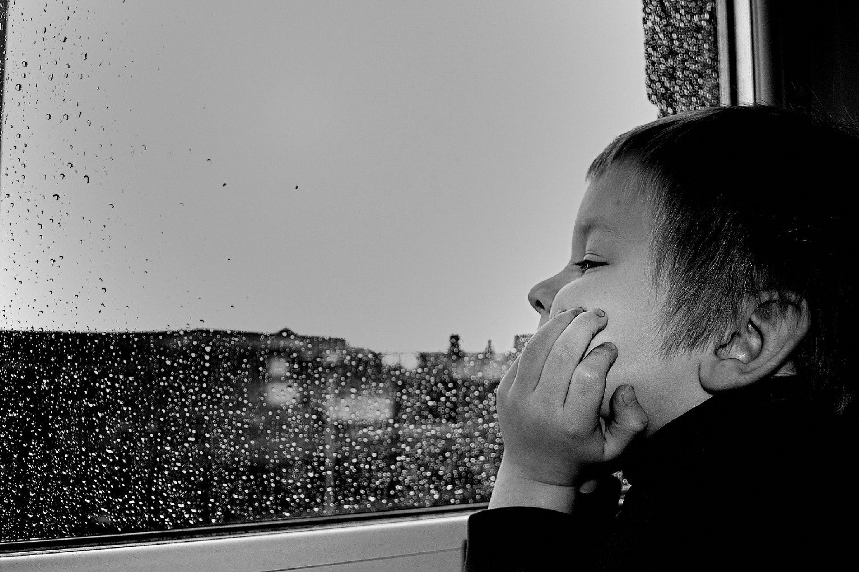 Ein Junge schaut aus einem Fenster. Draußen regnet es, Regentropfen auf der Scheibe.