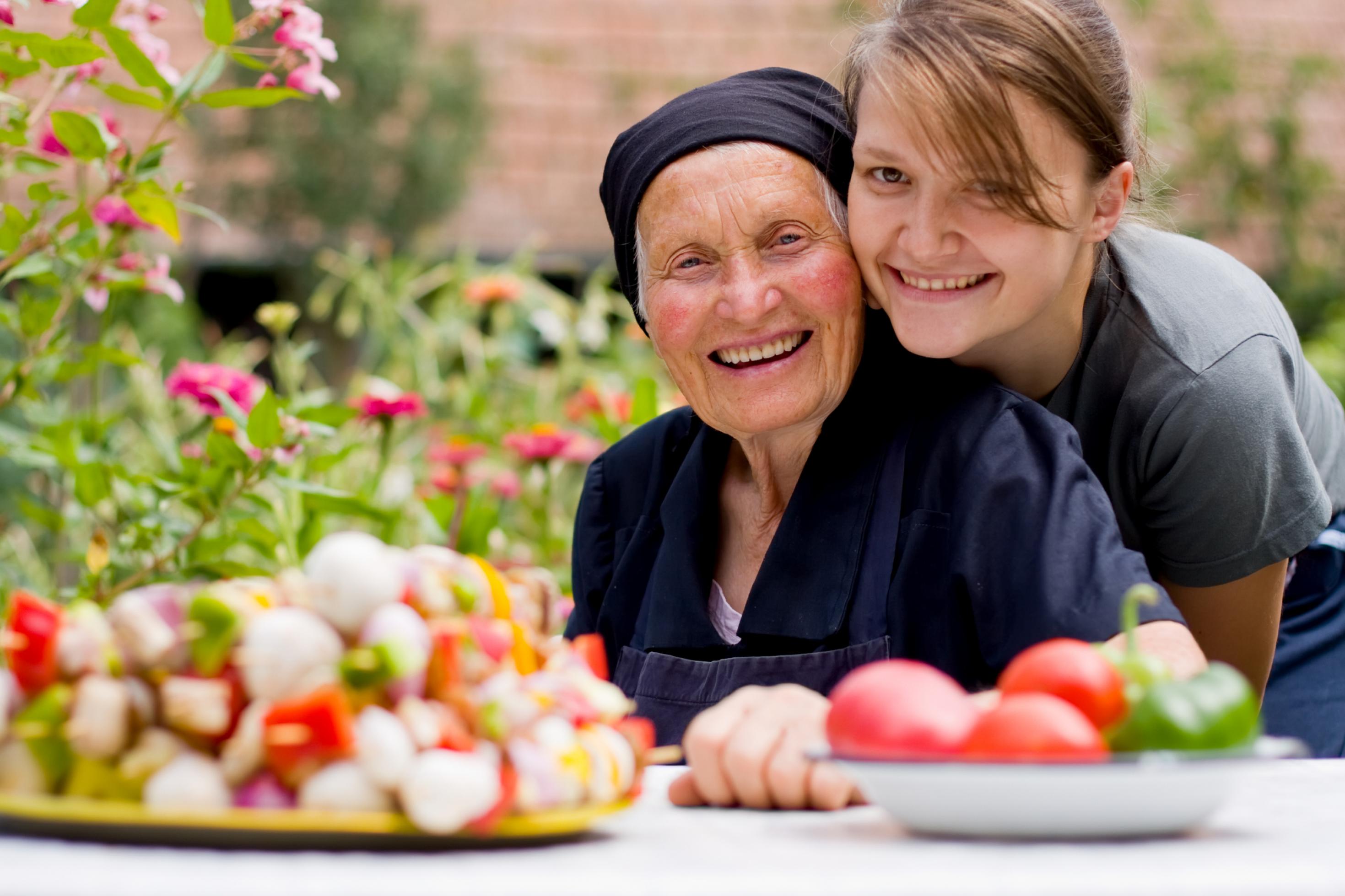 Eine junge Frau mit ihrer Großmutter an einem Tisch, auf dem zwei Teller mit frischem Gemüse stehen.