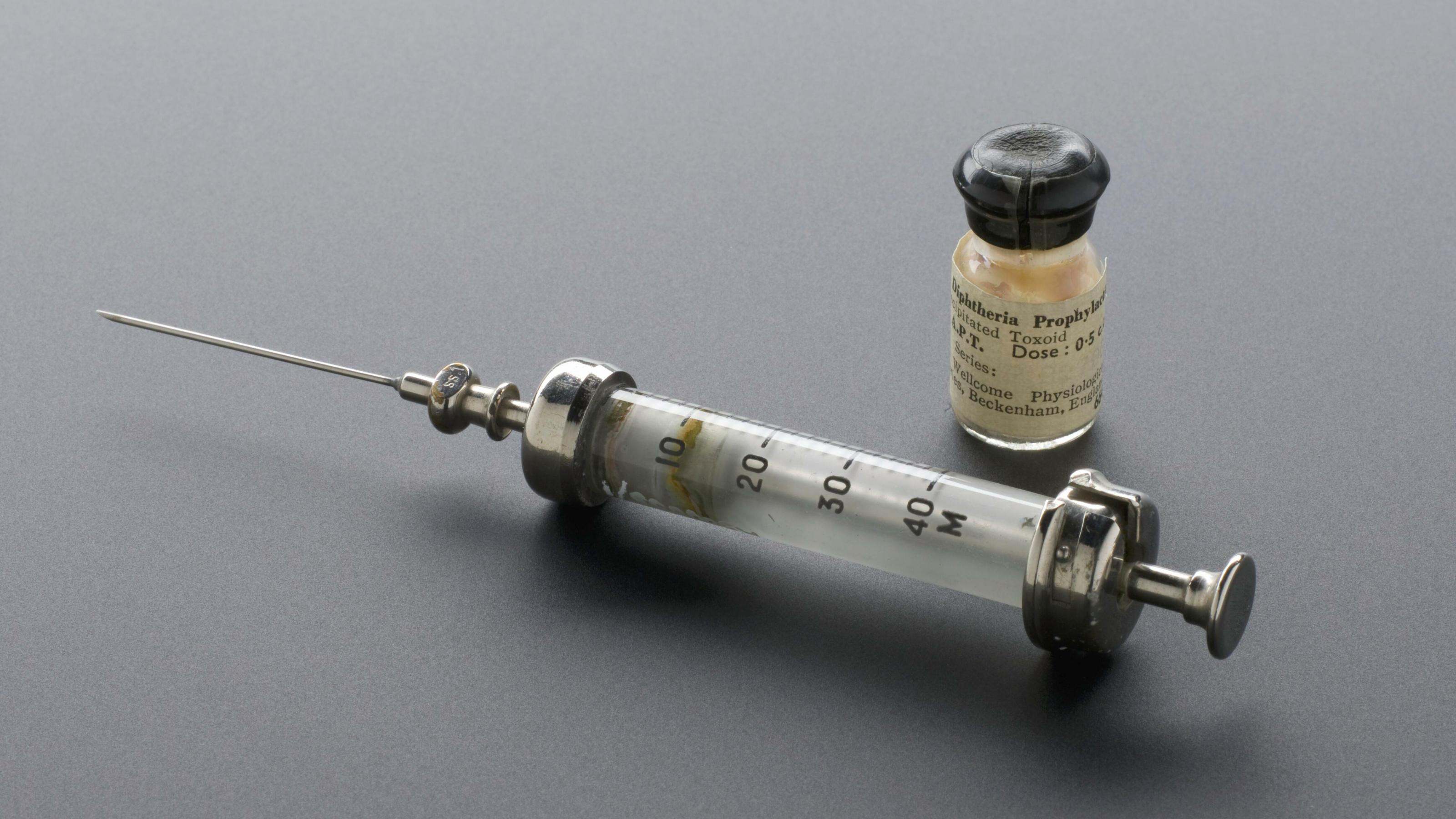 Auf dem Bild ist eine historische Impfspritze und ein kleines Fläschchen mit Diphtherie-Impfstoff zu sehen.