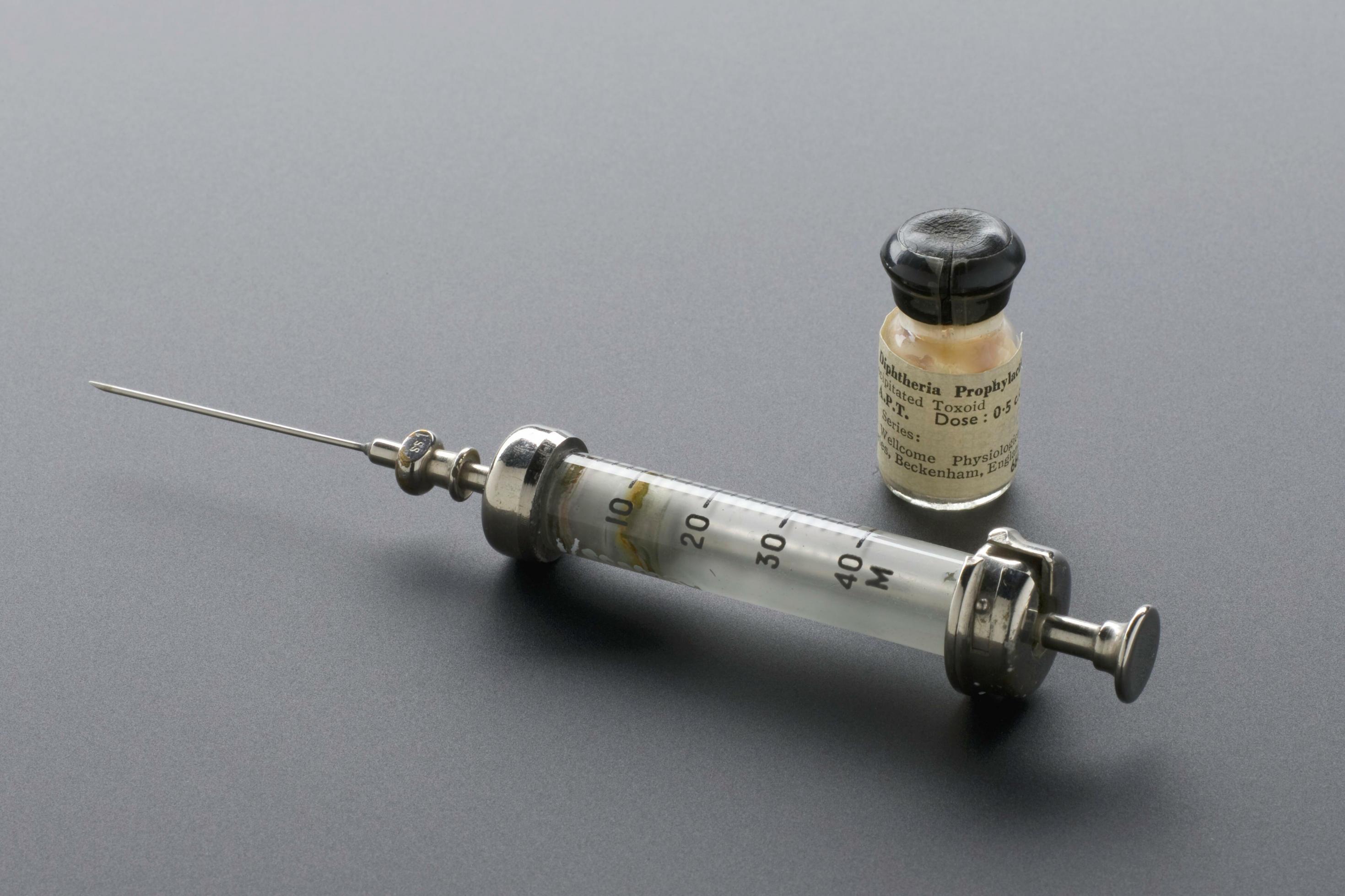 Auf dem Bild ist eine historische Impfspritze und ein kleines Fläschchen mit Diphtherie-Impfstoff zu sehen.