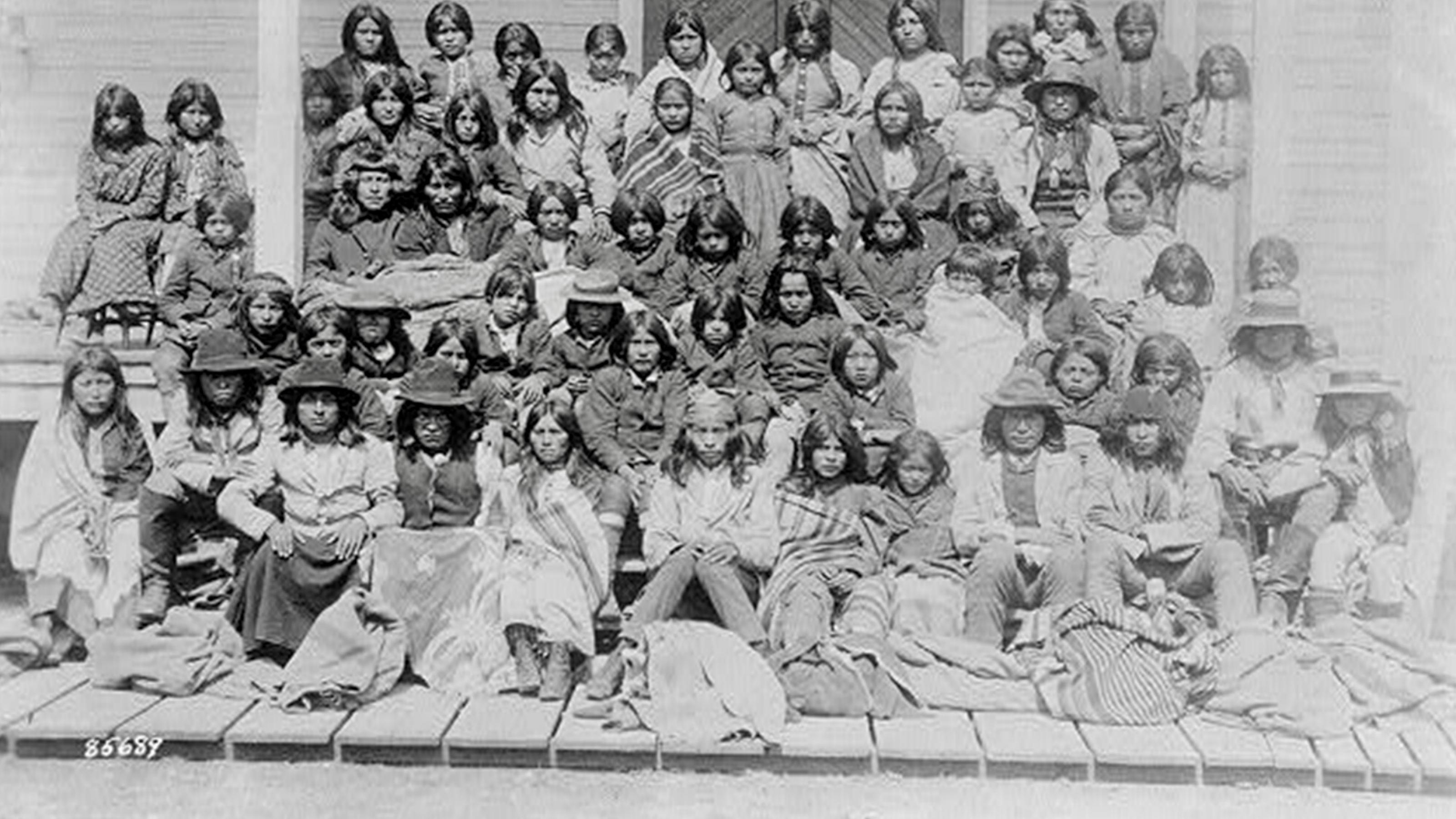 Eine Gruppe von etwa 50 Kindern posiert bei der Ankunft im Internat in Carlisle Pennsylvania in fünf Reihen sitzend auf den Treppen des Gebäudes. Die meisten haben langes dunkles Haar, manche tragen Decken über ihrer Kleidung. Sie schauen direkt in die Kamera