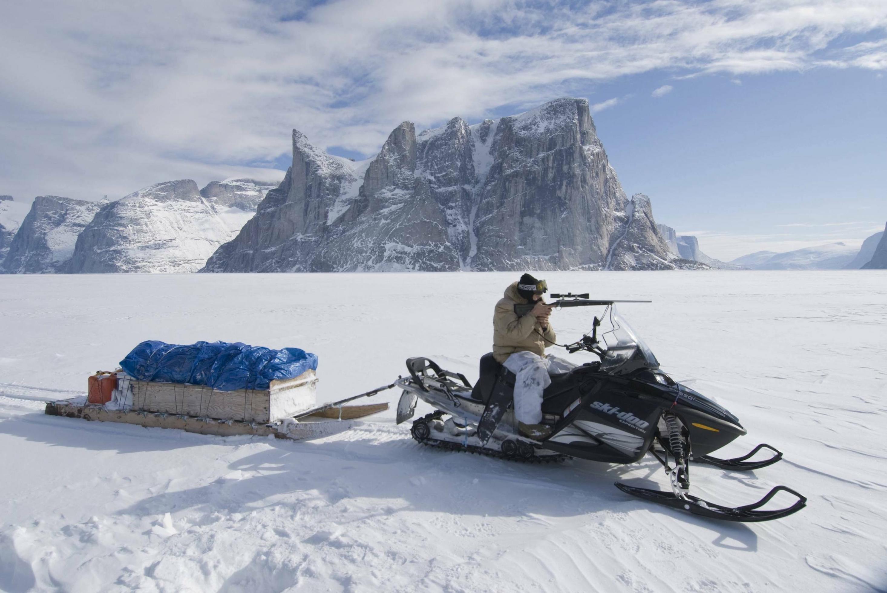 In einer schneebedeckten Landschaft mit hohen Bergen sitzt ein Inuit auf einem Motorschlitten und zielt mit einem Gewehr in die Ferne.
