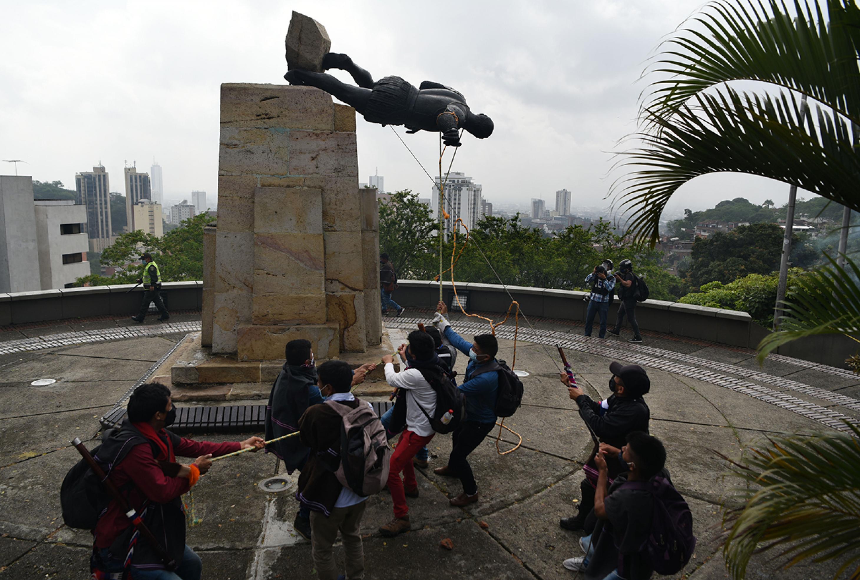 Acht Männer ziehen mit Seilen die Statue vom Sockel. Sie liegt waagerecht in der Luft. Im Hintergrund zwei filmende Männer und ein Polizist mit Schlagstock.