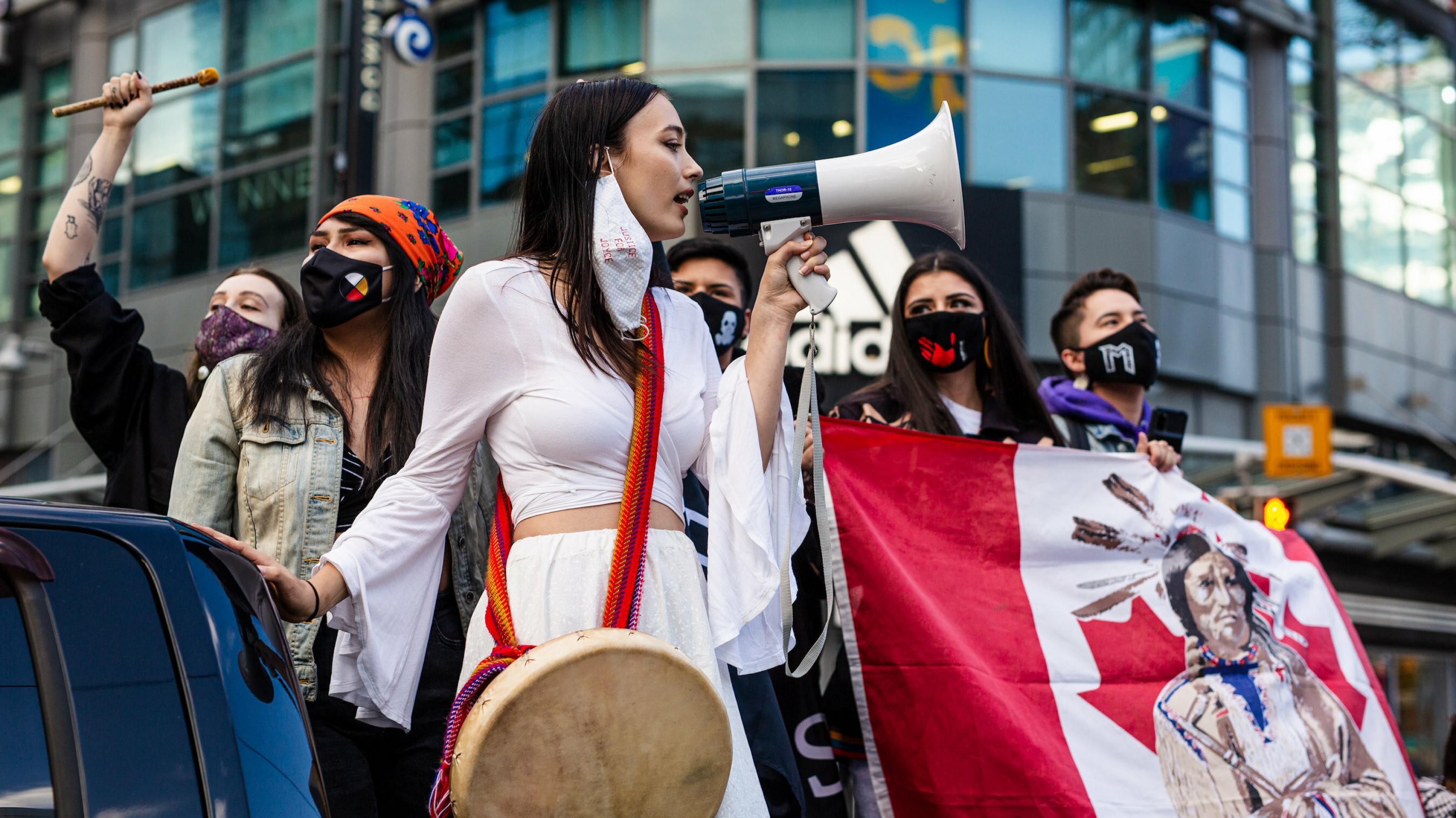 Junge Demonstrantinnen der kanadischen Indigenen stehen in einer Gruppe und halten ein Transparent mit der kanadischen Flagge, auf der ein indigener Häuptling zu sehen ist. Eine junge Frau in weißem Kleid spricht in ein Megafon.