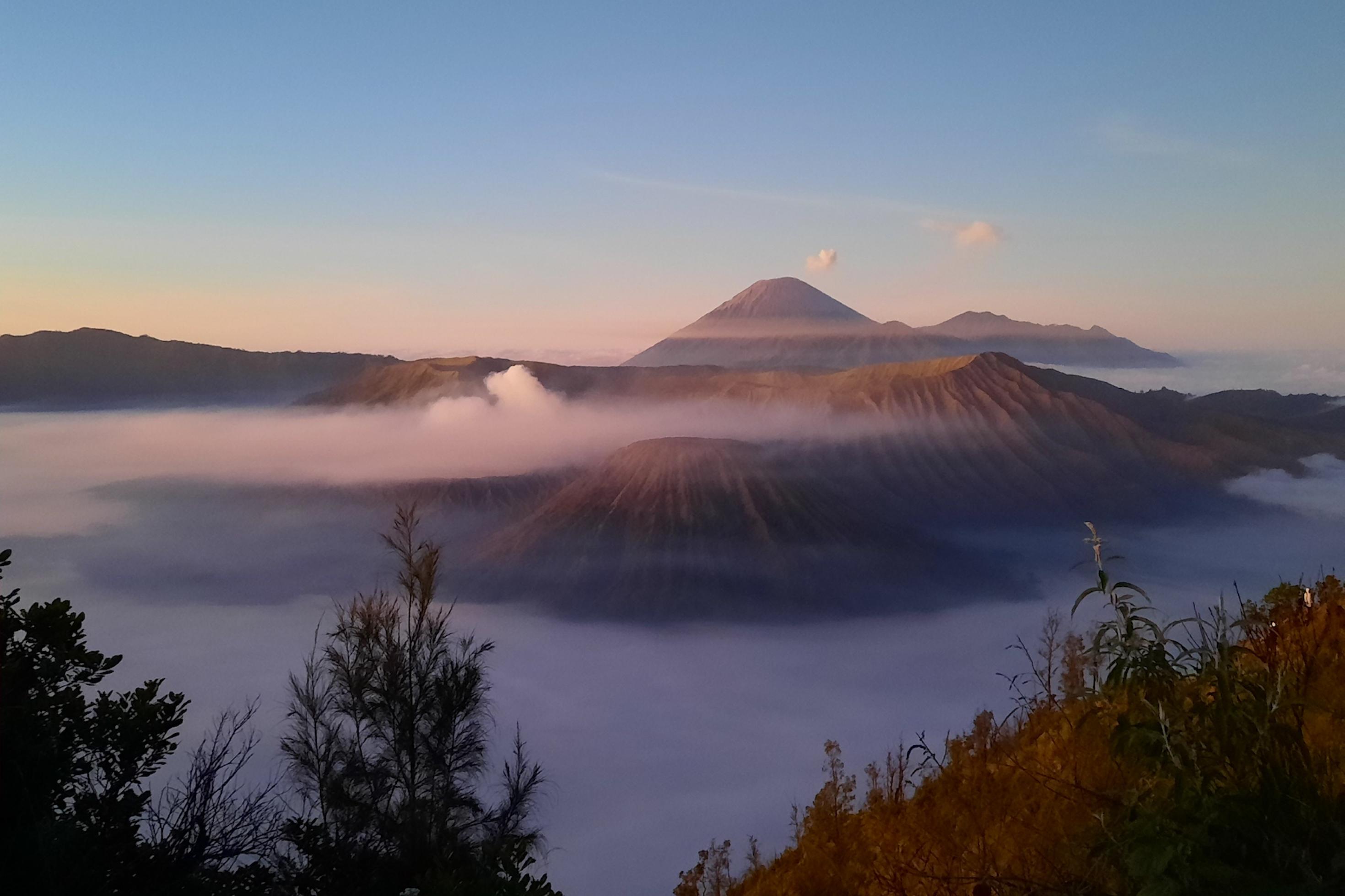 Blick auf den Krater der Vulkane Batok (vorne) und Bromo, die zum Sonnenaufgang aus der Wolkendecke herausragen. Dahinter erhebt sich Javas höchster Vulkan Semeru.