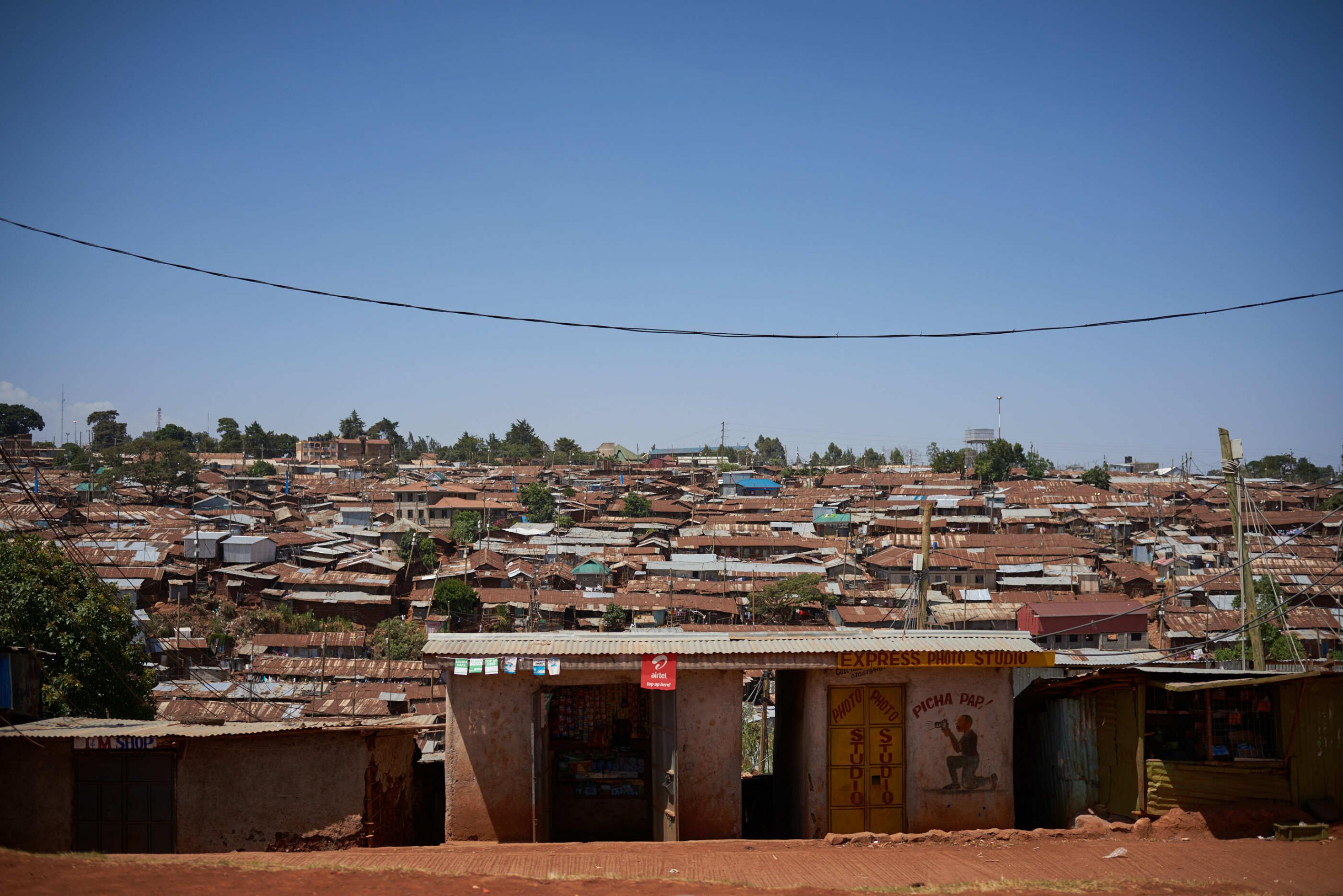 In einer Vorstadtsiedlung in Nairobi stehen Wellblechhütten. Man sieht nur ihre Dächer, die fast alle rostrot sind. Dazwischen schauen ab und zu kleine grüne Bäume hervor.