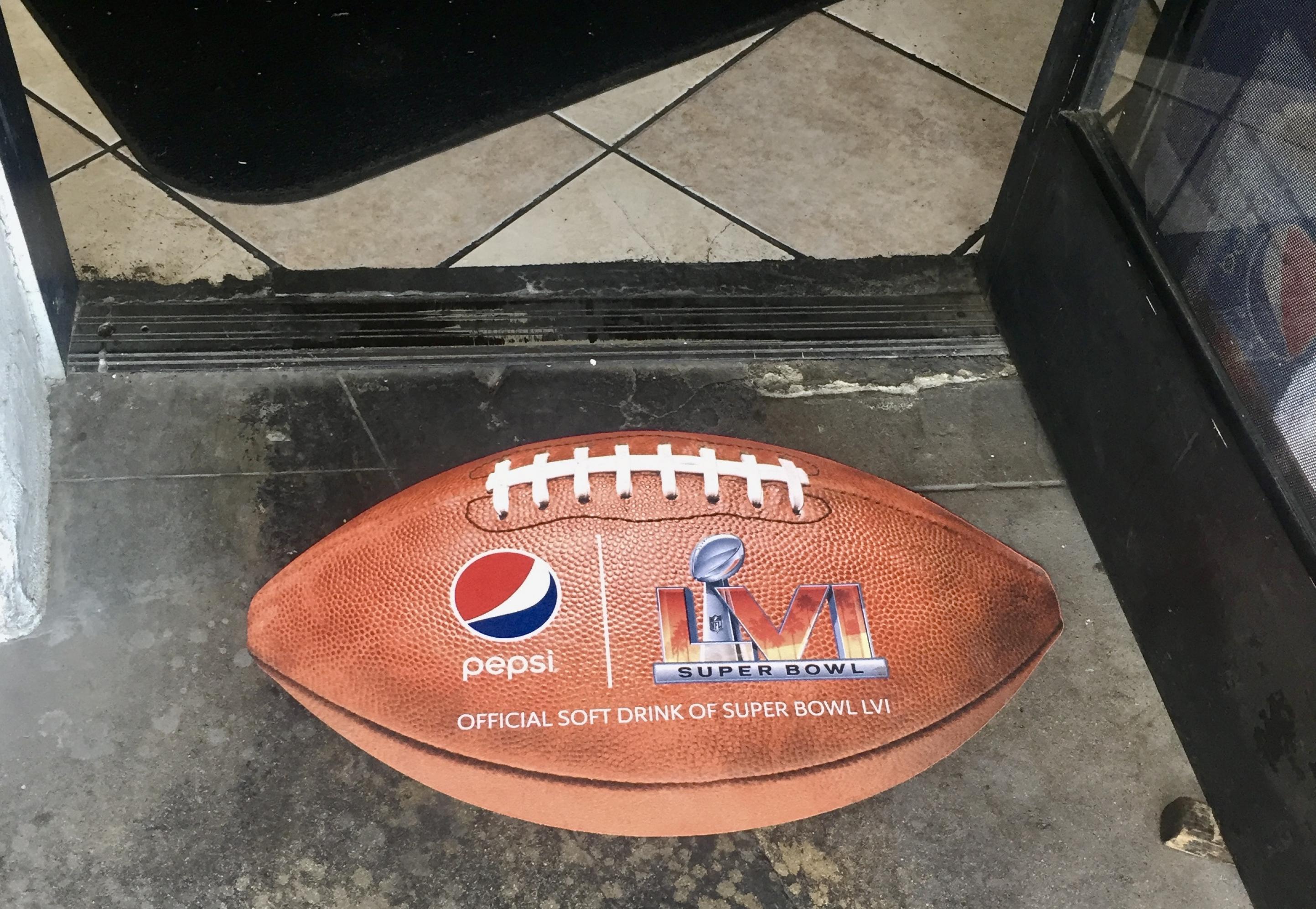 Ein Aufkleber in Form und Größe eines Footballs wirbt auf den Boden im Eingang des Imbiss restaurants Munchkins für den 56. Super Bowl und für den Sponsor Pepsi. Dahinter die kacheln des Fussbodens und eine schwarze Fußmatte.