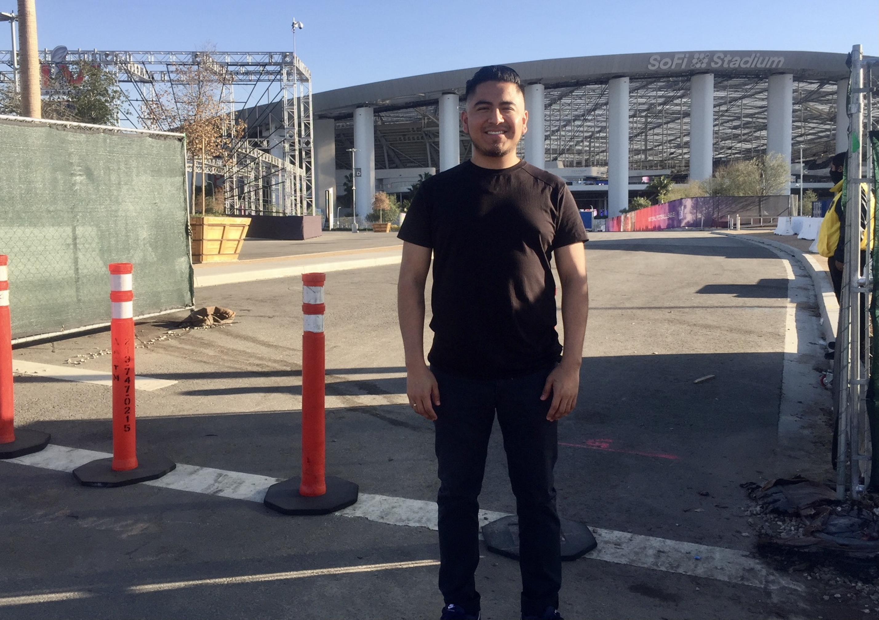 Óscar Alvarez, ein 27 Jahre alter Sozialarbeiter, posiert in schwarzen Jeans und schwarzem T-Shirt vor dem abgesperrten SoFi Stadion. Im Hintergrund ein Gerüst und Pflanzentöpfe.