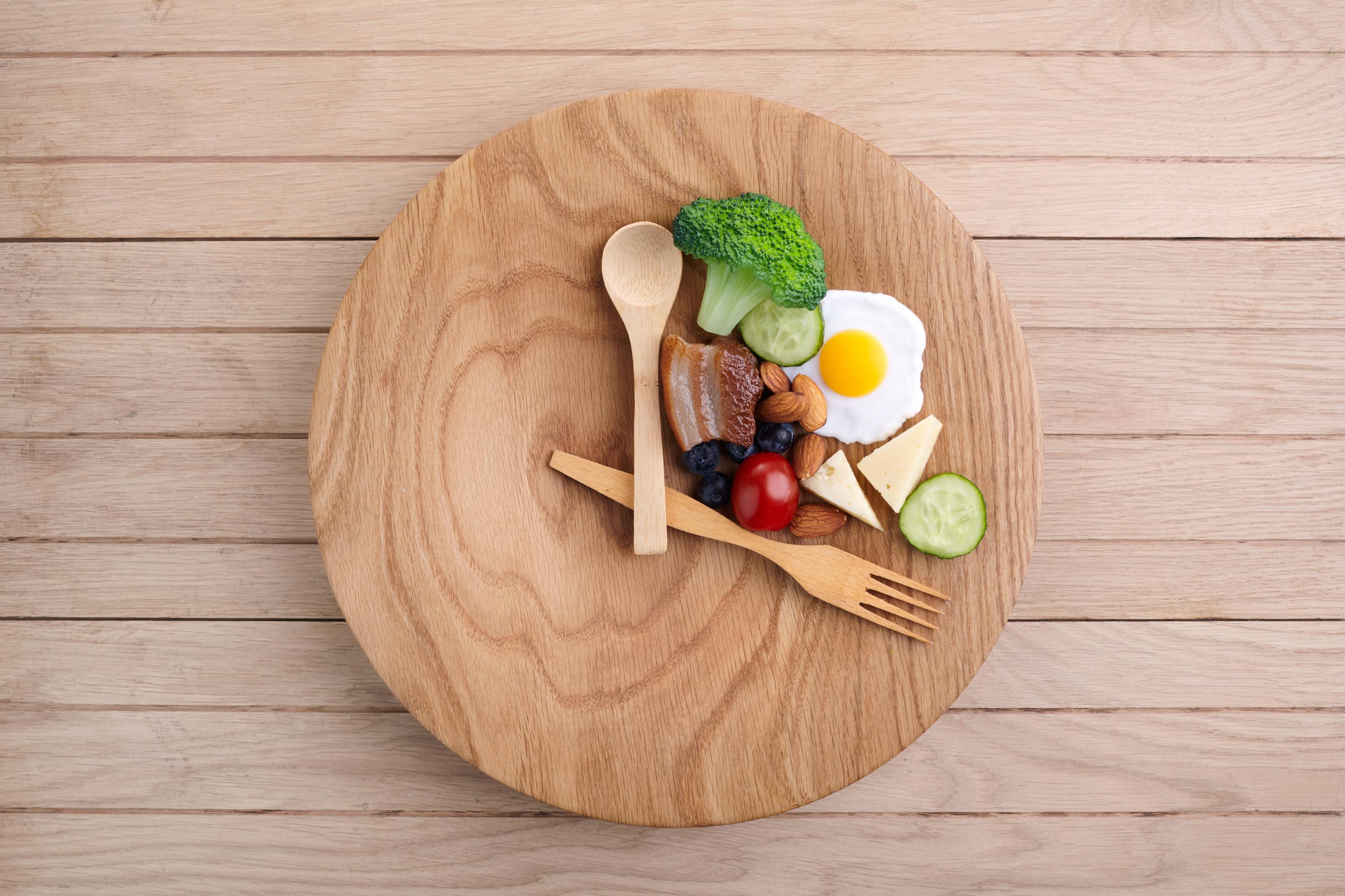Auf einem runden Holzteller sind Löffel und Gabel wie die Zeiger einer Uhr angeordnet. Oben rechts liegen verschiedene Lebensmittel.