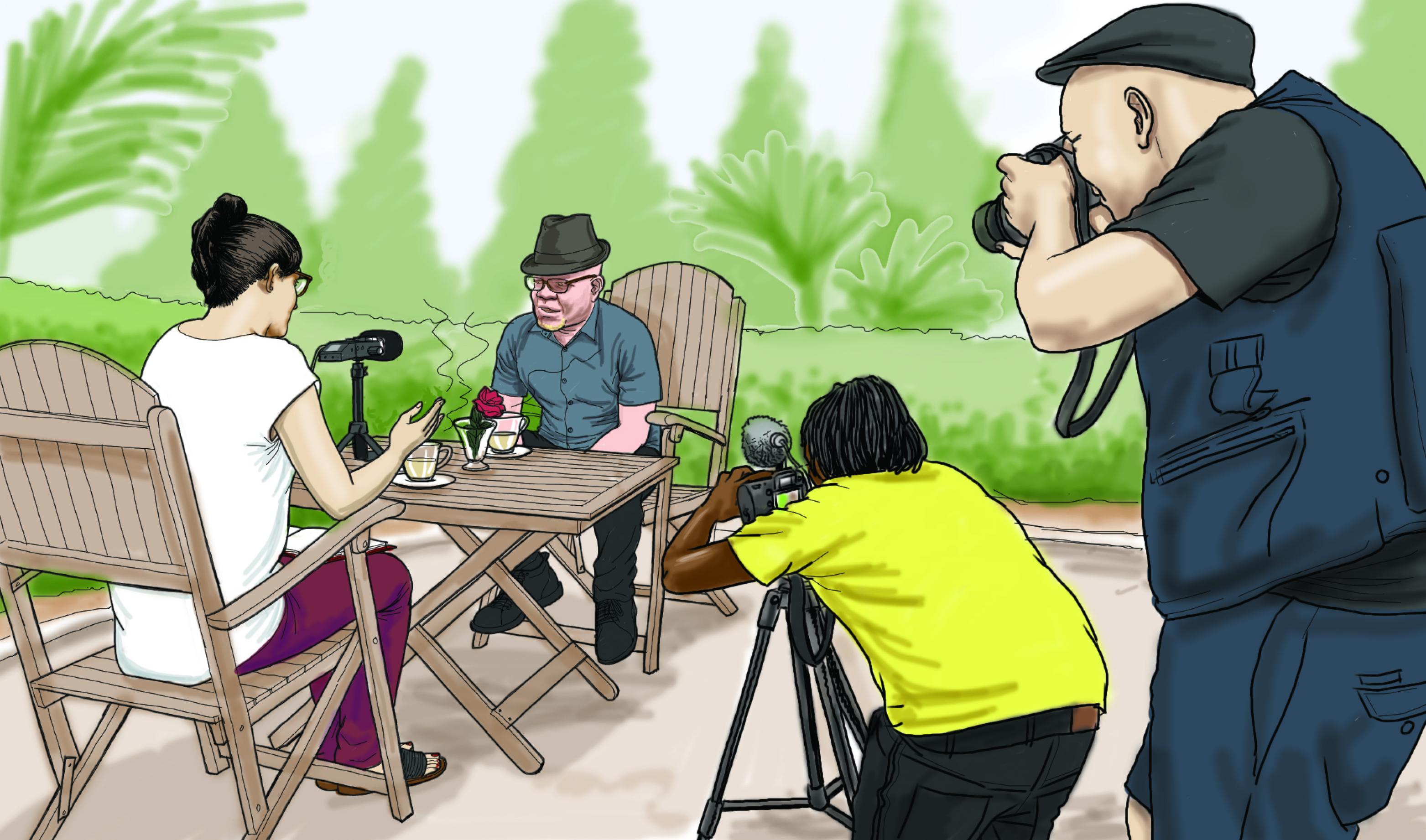 Eine weiße Frau und ein Mann mit Albinismus unterhalten sich in einer Interviewsituation. Im Vordergrund steht ein Schwarzer Mann hinter einer Filmkamera und ein weißer Mann fotografiert.