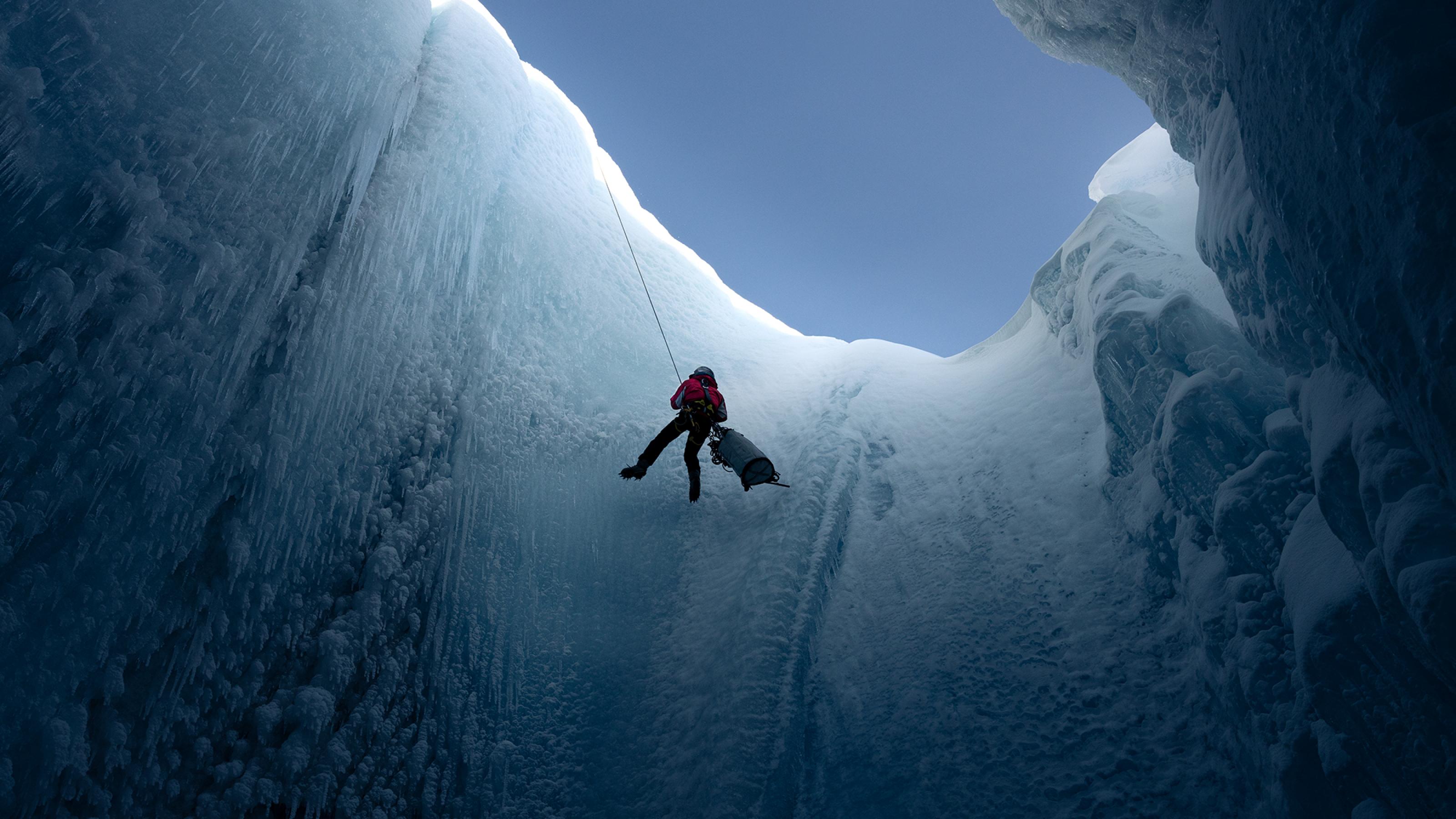 Aus der Tiefe einer Gletschermühle aufgenommen sieht man den Glaziologen Alun Hubbard am Seil hängend. Er ist gerade dabei, sich in das Eisloch im Gletscher abzuseilen.