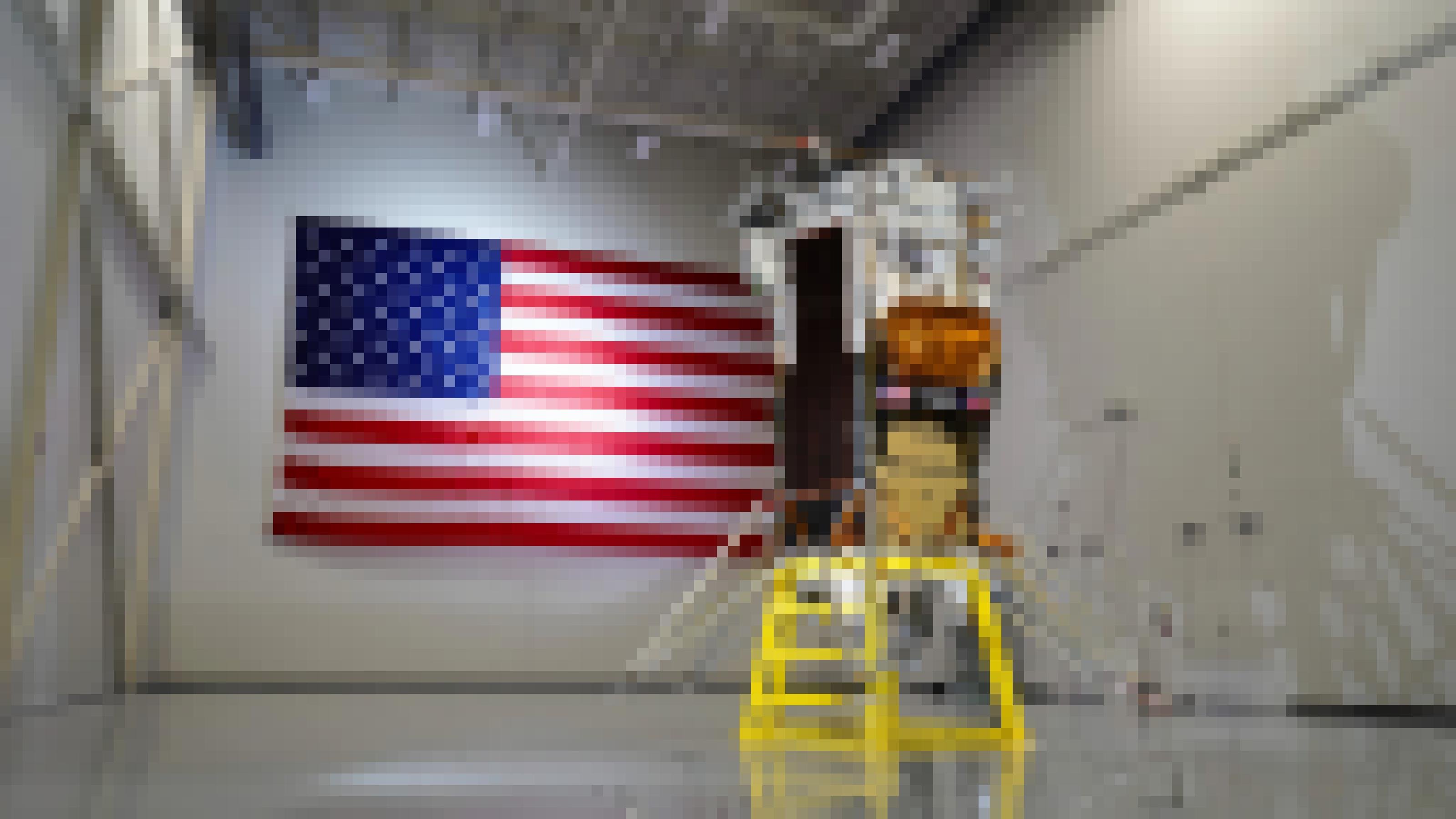 Eine Landefähre in einer leeren Lagerhalle. Im Hintergrund befindet sich eine riesengroße Flagge der USA.