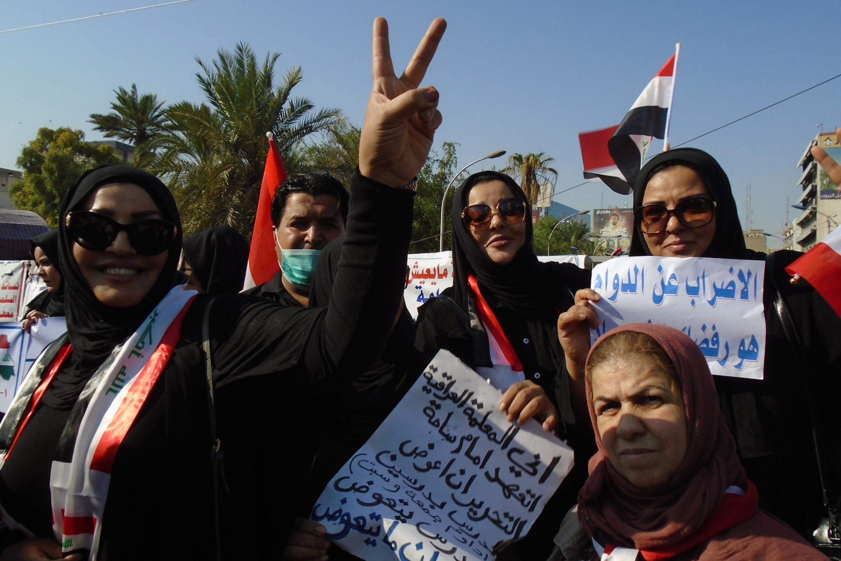 In der Mitte des Bildes ist eine hochgereckte Hand mit dem Siegeszeichen zu sehen, rechts davon die irakische Flagge, die offenbar ebenfalls eine Demonstrantin mitgebracht hat. Zwei von ihnen halten Demoschilder mit Parolen in arabischer Sprache in die Höhe.