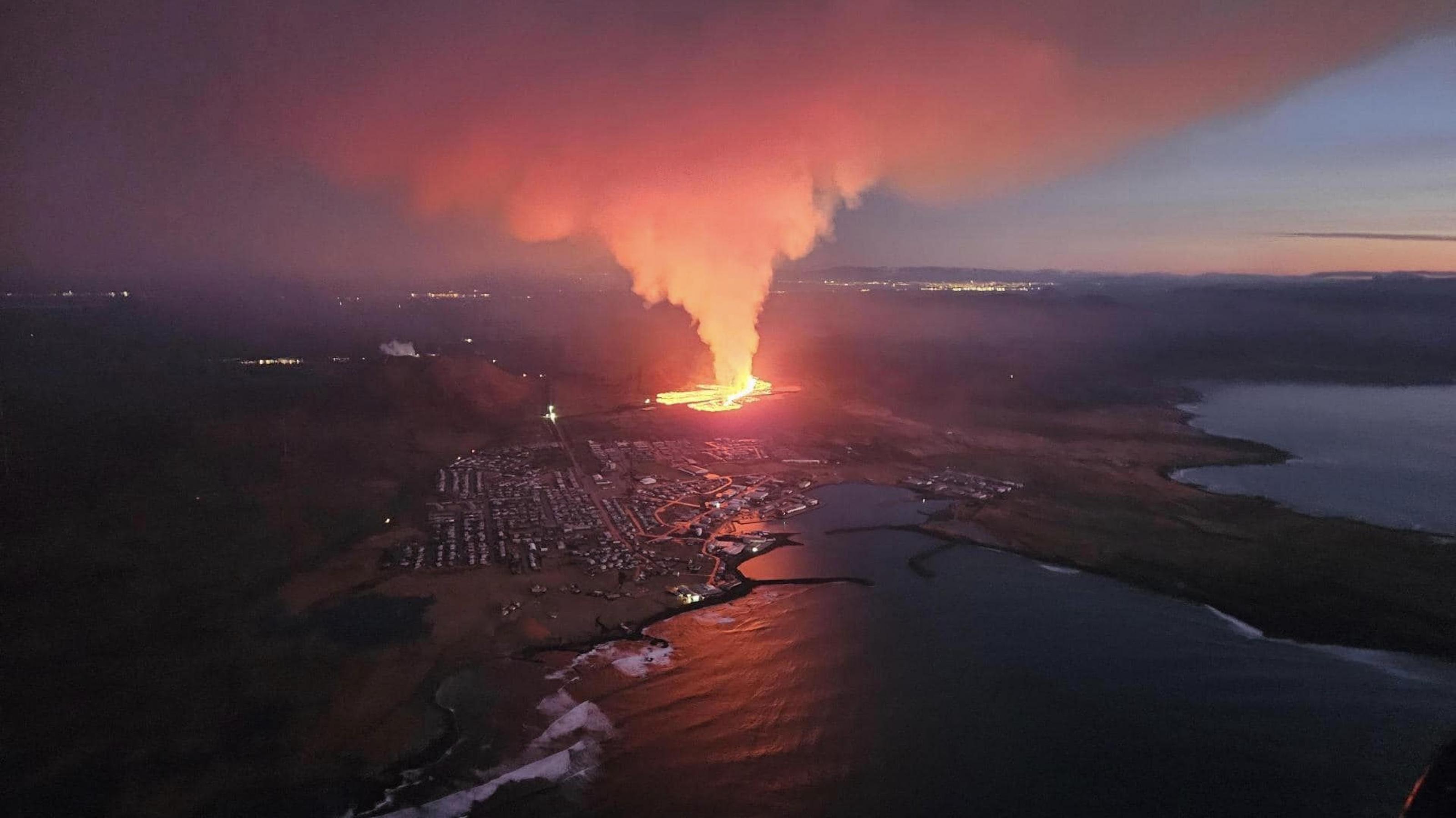 Luftbild, die noch fast dunkle Küste, der Ort Grindavik, dahinter erschreckend nah und gleißend hell der Lavastrom und eine große Rauchwolke.