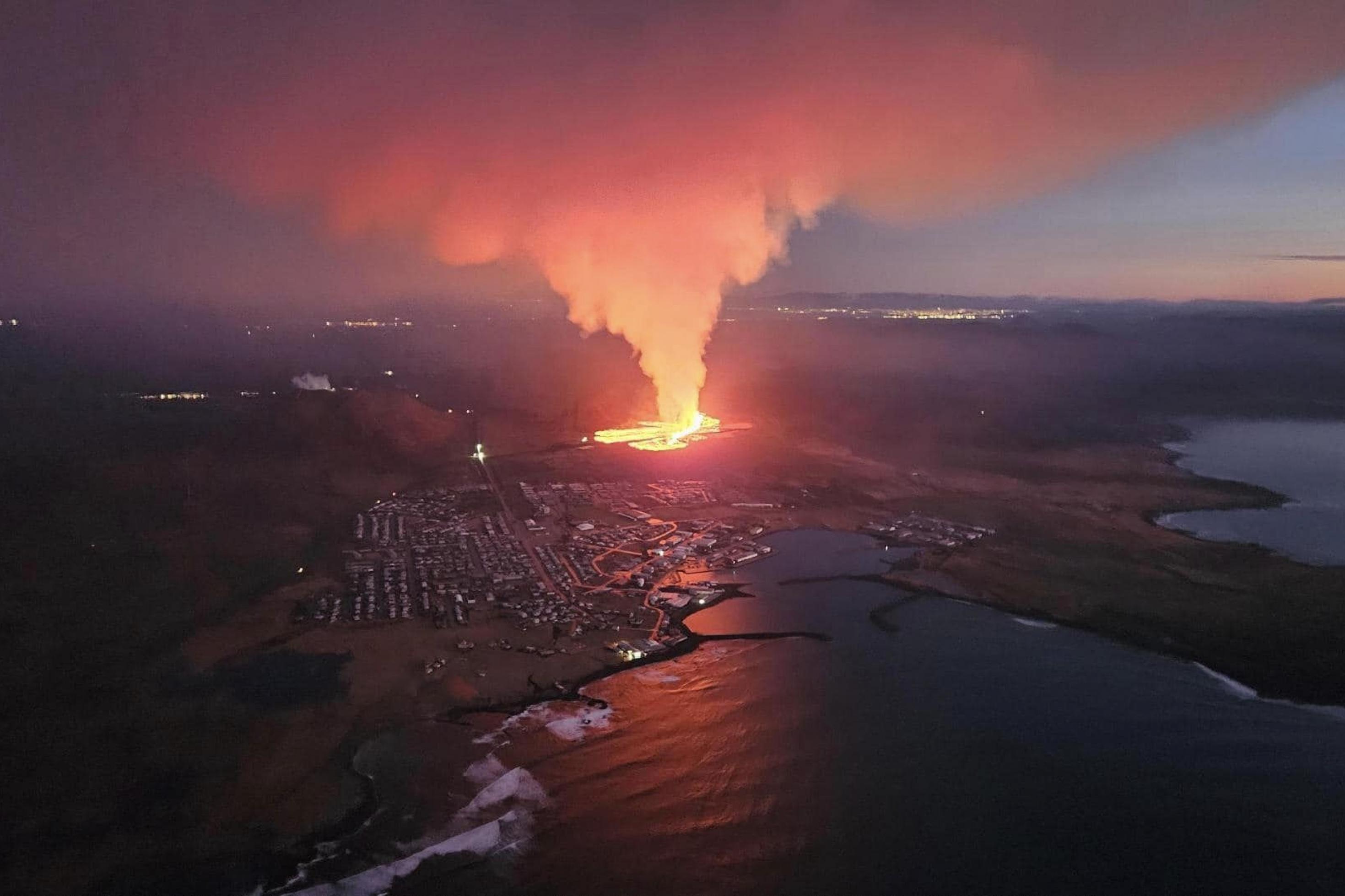 Luftbild, die noch fast dunkle Küste, der Ort Grindavik, dahinter erschreckend nah und gleißend hell der Lavastrom und eine große Rauchwolke.
