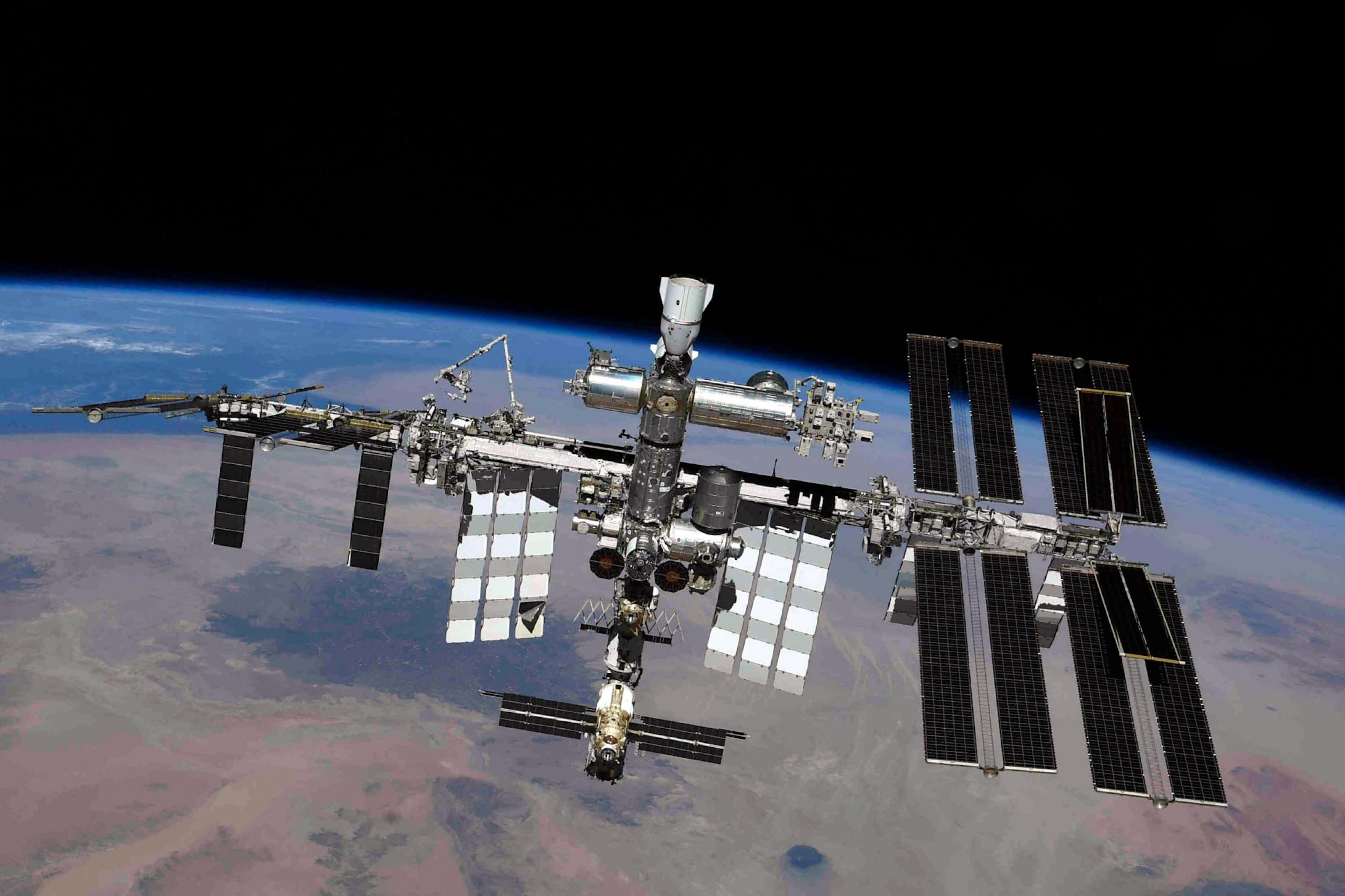Die ISS von außen, rechts mit den neuen, 2021 gelieferten Solarzellen, im Hintergrund die Erde mit bräunlichen Landmassen