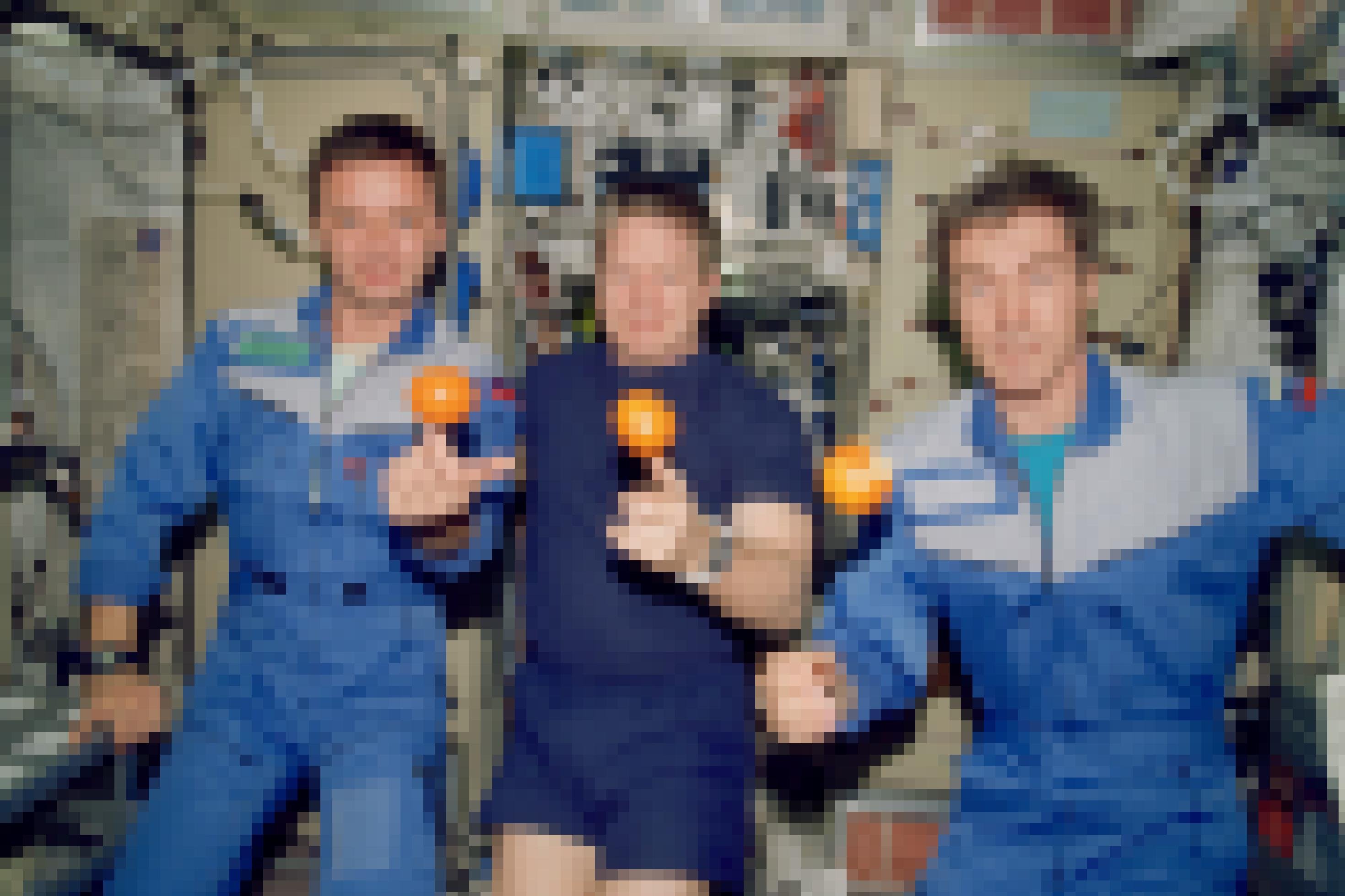 Drei leicht lächelnde Raumfahrer auf der Raumstation, über einer Hand von jedem schwebt eine Orange.
